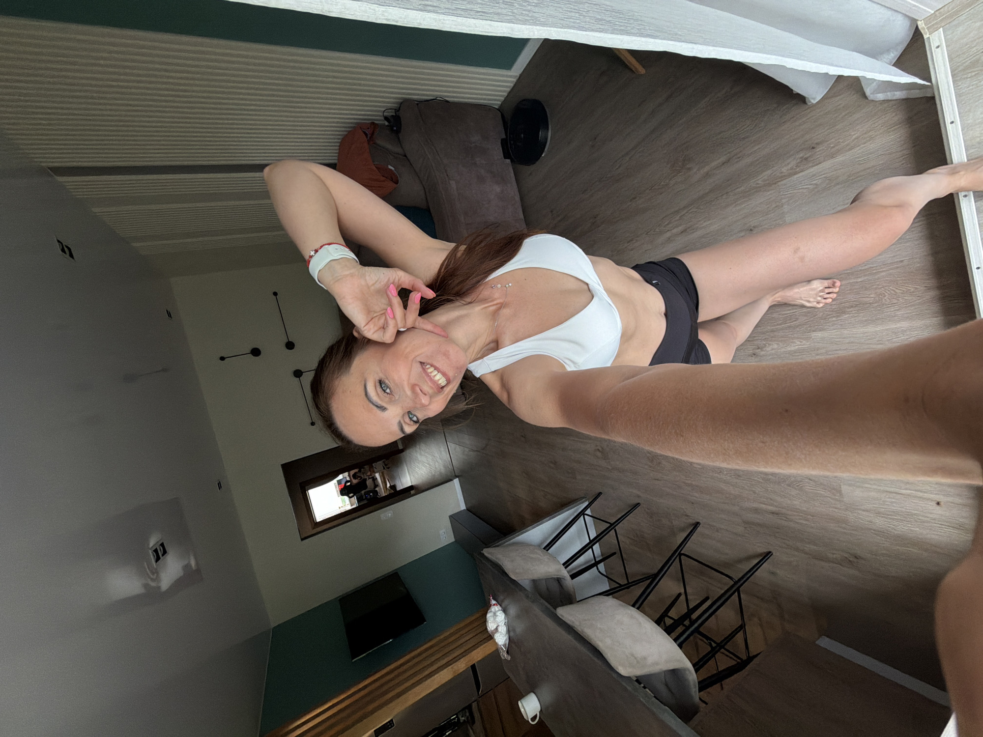 A woman is taking a selfie in a modern apartment. She is wearing a white sports bra and black athletic shorts. The woman has brown hair pulled back in a ponytail and is smiling at the camera, with her right arm raised and hand near her face. In the background, a dining table with chairs is visible, as is a doorway leading to another room. A robotic vacuum cleaner is on the floor in the distance. The apartment has light-colored wooden flooring and neutral-toned walls. The overall impression is a casual, domestic setting. This image does not contain any information that would place it in Birobidzhan, Russia.