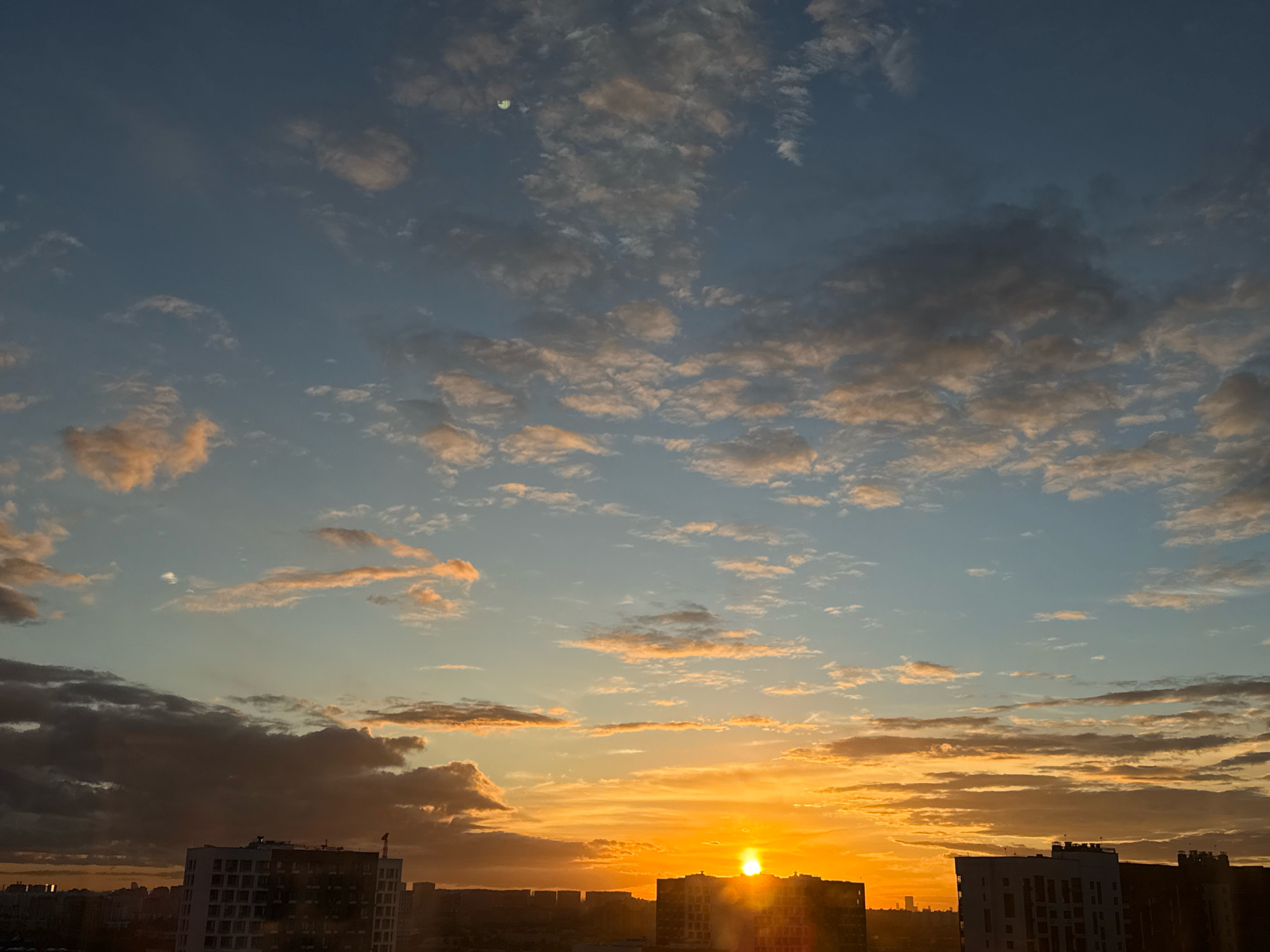 The image depicts a sunset over the city of Lyubertsy, Russia. The sky is filled with a gradient of colors, transitioning from a deep blue at the top to vibrant oranges and yellows near the horizon. Scattered clouds, some tinged with pink and gold from the setting sun, drift across the sky. In the upper left quadrant, a small, pale crescent moon is visible.
The lower portion of the image features a city skyline dominated by buildings. Several multi-story residential buildings are prominent, with their windows reflecting the light. The silhouette of other structures can be seen in the distance, suggesting a dense urban environment. The sun is low on the horizon, partially obscured by one of the buildings, casting a bright glow and creating lens flare effects. No people or specific actions are discernible in the image. The environment appears to be an urban setting at dusk.
