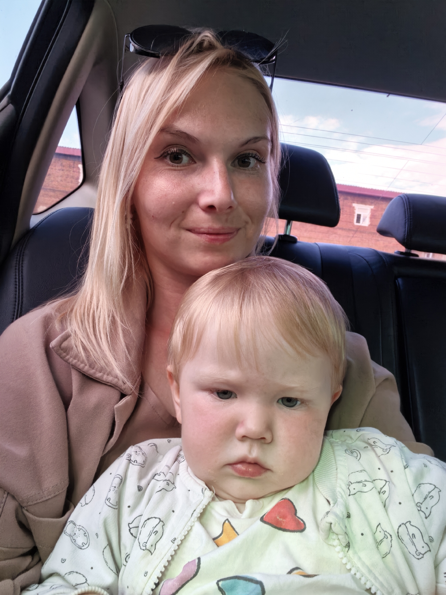 A blonde woman with sunglasses perched on her head is in the front passenger seat of a car, holding a baby. The woman is wearing a beige jacket. The baby, also with blonde hair, is wearing a white onesie with a pattern of cartoon animals and hearts. They are both looking at the camera. The background shows the interior of a car, including black leather headrests, and through the windows, a brick building and a cloudy sky. The context of Sterlitamak, Russia is not visually determinable from the image.