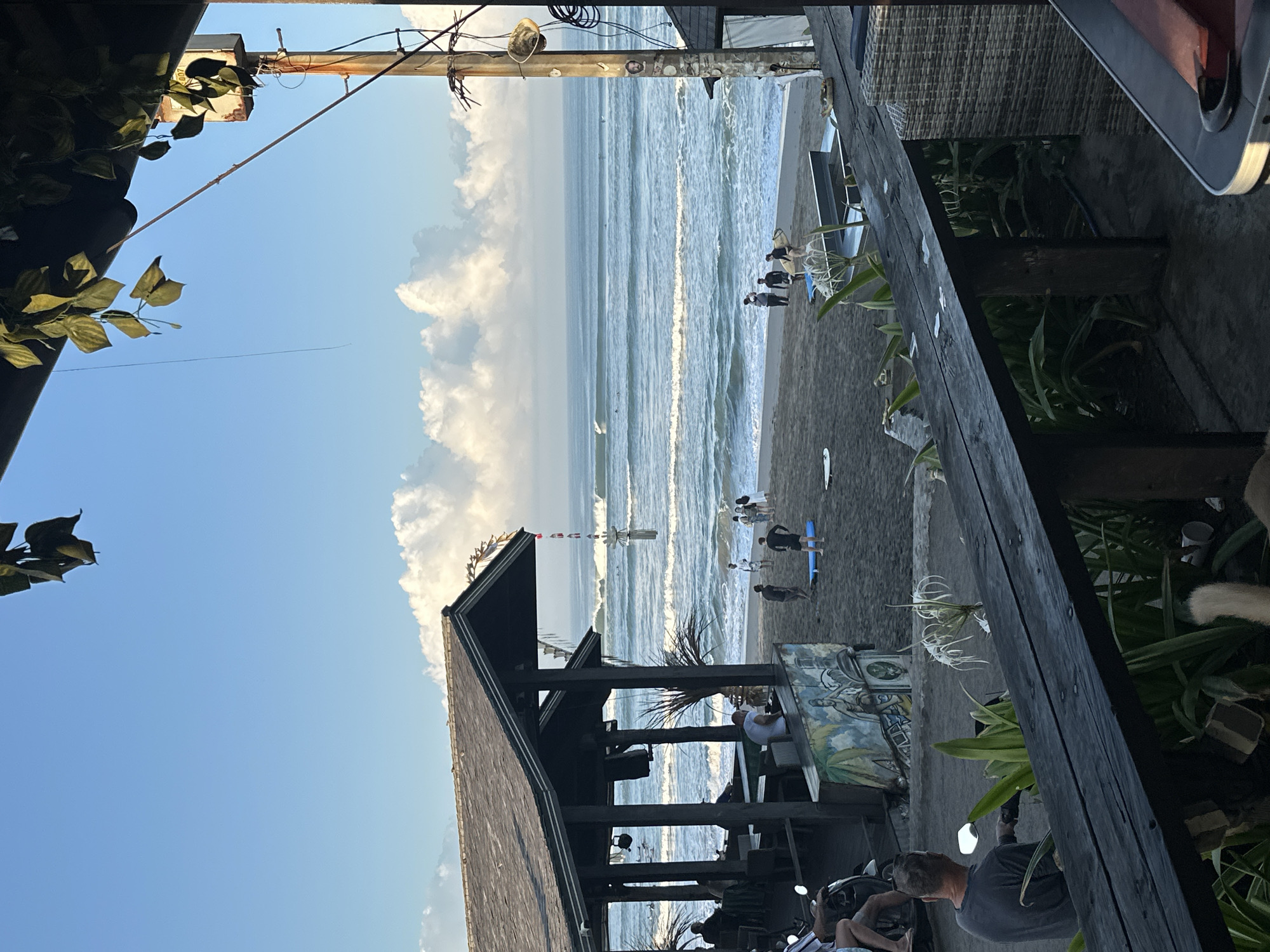 The image depicts a beach scene in North Kuta, Indonesia. In the foreground, a dark wooden railing and a wicker chair are visible, suggesting an outdoor seating area of a restaurant or cafe. Beyond this, the sandy beach is populated by several individuals, some carrying surfboards, indicating a surf spot. The ocean features moderate waves, and a few surfers can be seen in the water. In the background, a open-air structure with a dark roof and pillars, likely a beach bar or restaurant, is present. The sky is blue with scattered white clouds. The overall impression is a relaxed, tropical atmosphere.