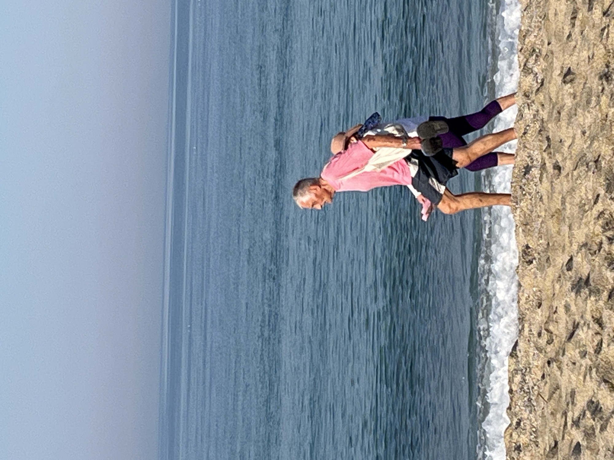 The image captures a man giving a piggyback ride to a woman on a sandy beach. The man wears a pink shirt and dark shorts, while the woman has on a white shirt, purple leggings, and dark shoes. They are positioned near the water's edge where waves are breaking. The backdrop consists of the ocean and sky. While the exact location is difficult to determine solely from the image, the city of Alanya, Turkey, is known for its beaches and coastline, suggesting this could be a coastal area there.