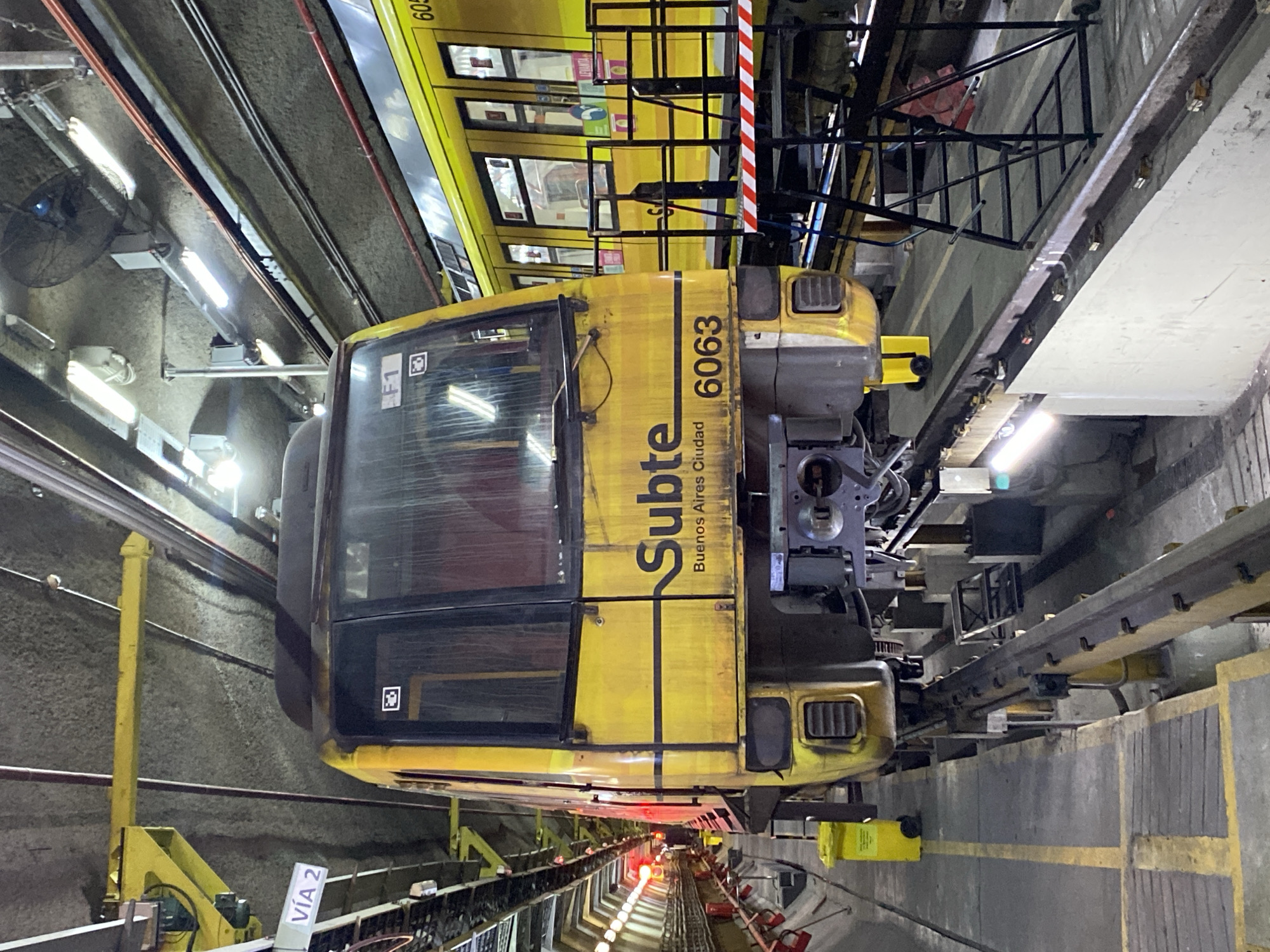 The image captures a yellow train car in an underground or tunnel environment, likely part of the Buenos Aires subway system, known as "Subte". The perspective is slightly elevated, looking towards the front of the train and down the tracks.
The primary subject is a yellow train, which appears to be a specialized service or maintenance vehicle due to its distinct, somewhat open-front cabin design and visible machinery at the front. The word "Subte" is prominently displayed vertically on its side in black, followed by "Buenos Aires Ciudad" and the number "6063". The front of the car shows signs of wear and features grilles and technical equipment. Below the cabin, various mechanical components and structural elements are visible. Further down the tracks, several red lights are illuminated, suggesting signaling or the end of a track section.
In the upper background, another yellow train car, partially visible, appears to be a standard passenger car with windows. The setting is an industrial tunnel, characterized by concrete walls, overhead fluorescent light fixtures, ceiling-mounted fans, and a network of electrical conduits and pipes. Black metal support structures or guardrails run alongside the tracks. Near the bottom left, a white sign clearly displays "VIA 2" (Track 2), indicating track identification. The entire scene is illuminated by artificial lighting, consistent with an indoor, operational area such as a maintenance depot or an underground line. No people are visible in the image.