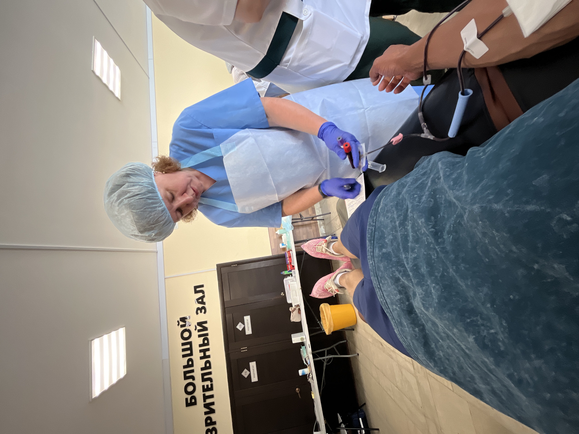 The scene depicts a blood donation process, possibly taking place in the city of Chaikovsky, Russia, based on the inscription "БОЛЬШОЙ ЗРИТЕЛЬНЫЙ ЗАЛ" which translates to "Large Auditorium Hall".
A healthcare worker wearing a blue scrub suit, hair net, and blue gloves is interacting with a blood sample tube. The person receiving the blood donation is visible from the chest down, wearing blue clothing and pink shoes.
The blood draw is in progress, with tubing visible connected to the person's arm. Another individual in a white lab coat is partially visible standing nearby.
The room has light-colored walls and ceiling with rectangular light fixtures. There is a dark brown cabinet along the wall and a table with various medical supplies. A yellow waste container is also present.
