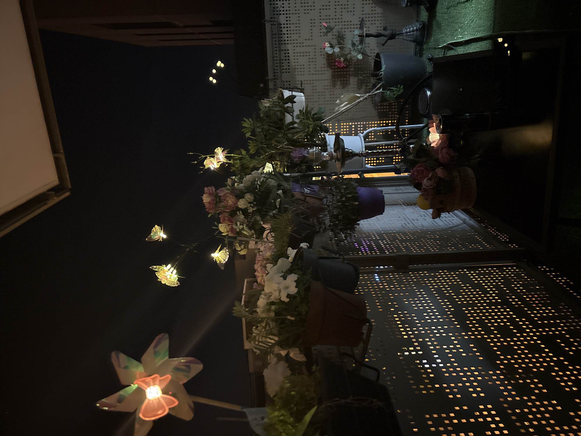The image is an indoor or outdoor shot taken at night in what seems to be a cafe or restaurant, potentially in Strasbourg, France. The foreground contains various decorative elements including faux flowers and illuminated fixtures such as a flower-shaped light. Tables and chairs suggest a place of dining and possibly outdoor seating. The presence of potted plants and ornate metal latticework indicates a deliberate attempt at creating an aesthetic or decorative environment. There is no evidence of people present in the frame.