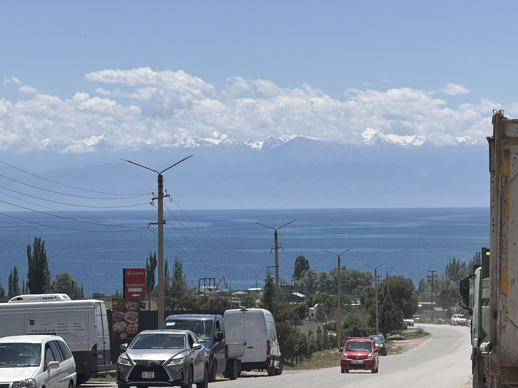 This image depicts a roadside scene with a body of water and distant mountains under a bright, cloudy sky. In the foreground, several vehicles are present, including a silver Lexus SUV with a license plate reading "01 978 AOD," a white delivery van with text that includes "Kerstner" and a phone number, and a red car. A large truck occupies the right side of the frame. Power lines crisscross the sky, supported by utility poles, some of which are topped with modern streetlights.
Behind the road, a wide expanse of blue water stretches towards a mountain range. The mountains are snow-capped, suggesting a significant elevation. A red sign with the text "db Demir Bark" and "БАНКОМАТ" (ATM in Russian) is visible, indicating a banking service. The overall scene suggests a travel or transportation setting, possibly in a region with both natural beauty and some level of development. The time of day appears to be midday, given the strong sunlight and the clear visibility. The weather is partly cloudy, with large white clouds contrasting against the blue sky. There are no people prominently visible in the image. The setting appears to be a somewhat rural or semi-urban area along a major road, with the natural landscape serving as a backdrop.
