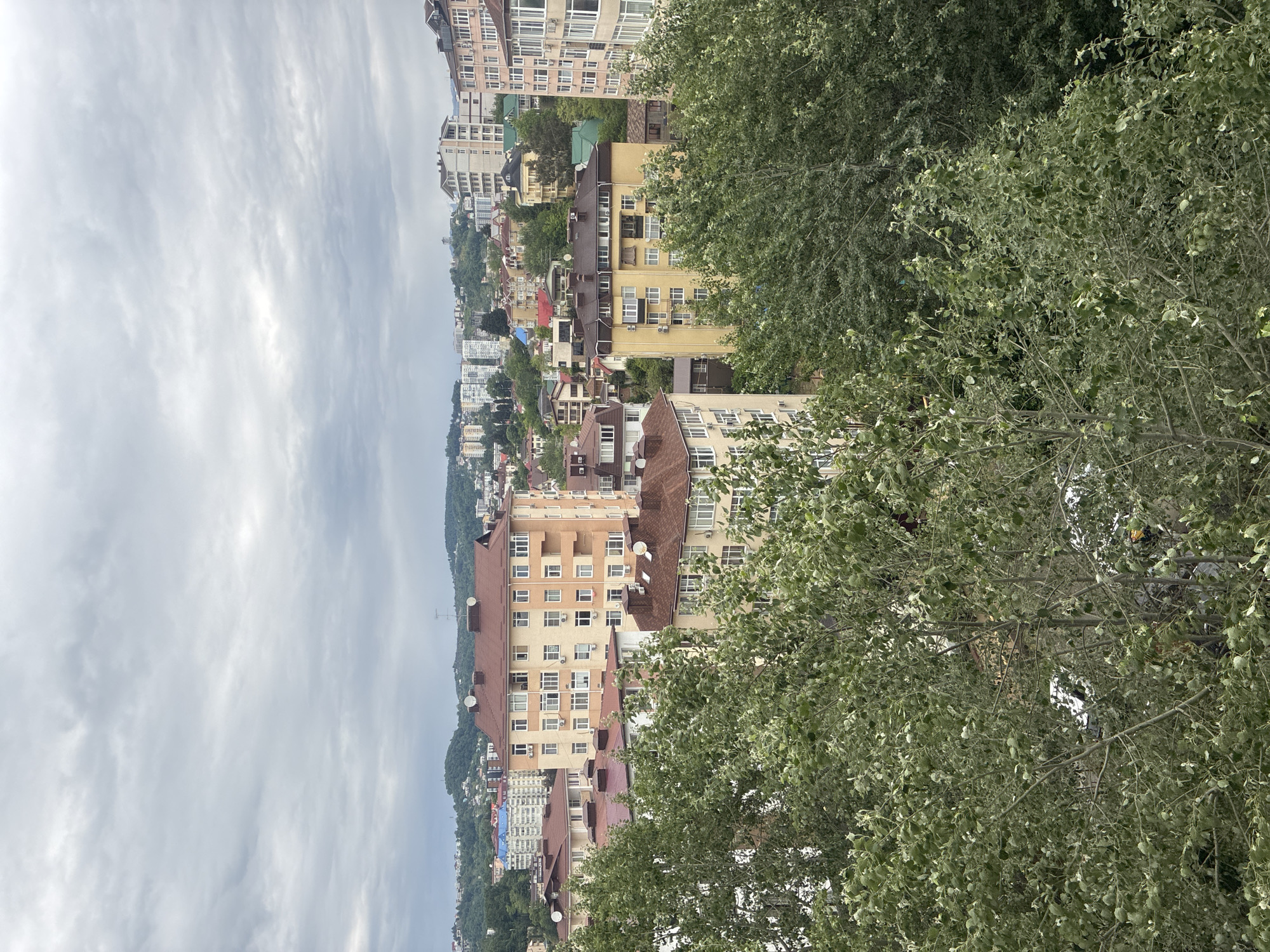 This is an outdoor scene in Sochi, Russia, showing a cityscape viewed from an elevated position. The sky is overcast with a layer of striated clouds. In the foreground, there are trees with green leaves filling the right side of the frame. The city itself consists of residential buildings of varying heights and colors. Some buildings have red tile roofs, while others are modern apartments with light-colored facades. There is a hillside visible with scattered greenery and more buildings nestled among the trees. The city is on a slope leading down towards what appears to be a coastline. Satellite dishes can be seen on top of some of the buildings. No people are visible in the scene.