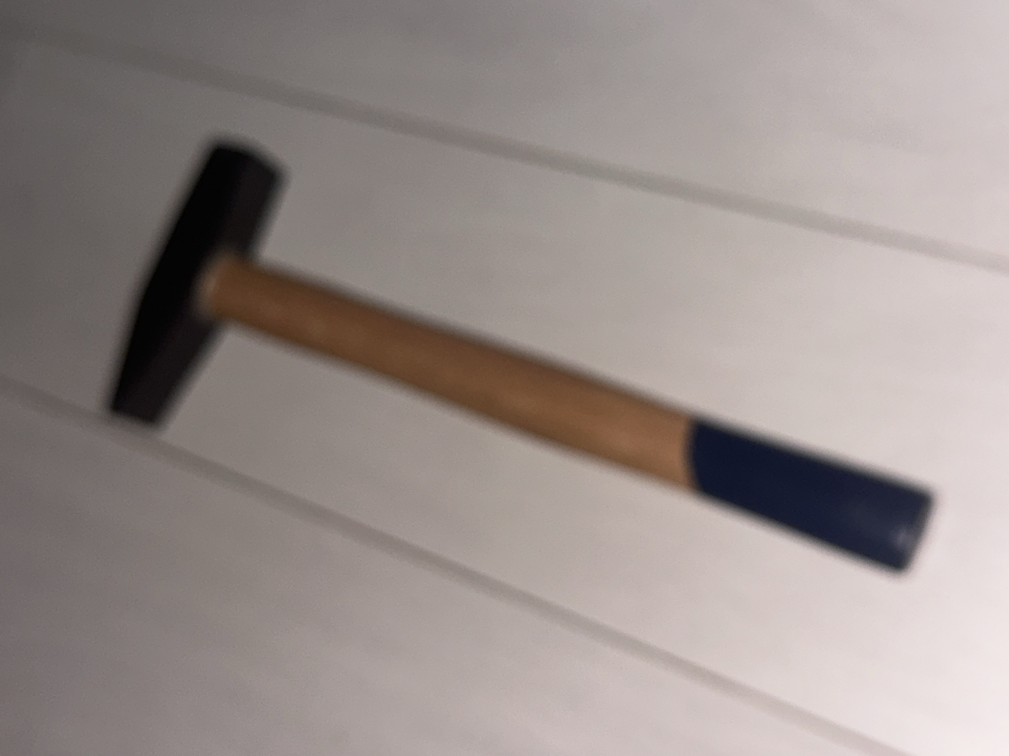 A hammer with a dark head and a wooden handle with a dark blue grip is positioned diagonally on a light-colored, possibly white, surface. The surface appears to be a floor or a table with faint lines suggesting planks or seams. The hammer is out of focus, indicating a blurred image. There are no people or any other objects visible in the frame. The scene is indoors, but the specific location within Sofia, Bulgaria, cannot be determined from the provided image.