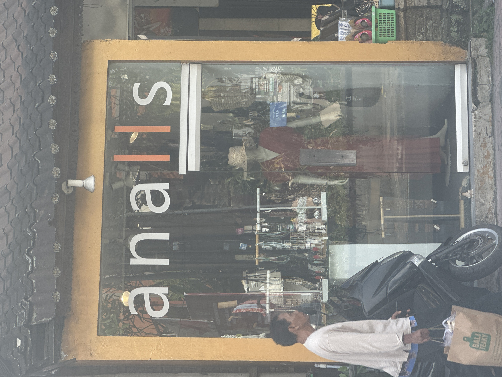In Ubud District, Indonesia, a shop displays "analis" in large white lettering across its storefront window. The window reveals mannequins wearing clothing and hats, along with racks of jewelry, bags, and other retail items. To the left, a man walks by holding a brown paper bag labeled "BALI TEAKY" and stands next to a black scooter. A green plastic basket containing footwear and other goods is visible outside the shop to the right. The shop has an orange-colored facade.