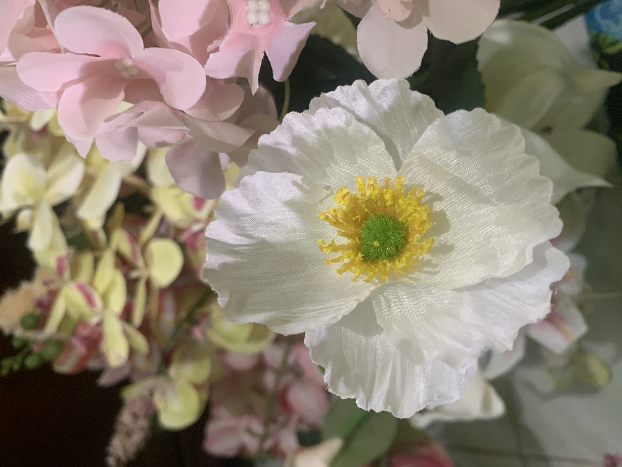 A close-up shot features a white flower with delicate, ruffled petals and a vibrant green center surrounded by yellow stamens. The flower is part of a bouquet that includes pink and yellow flowers, possibly artificial due to the texture and uniform coloring. The background is dark and blurred, suggesting a dimly lit indoor setting. The image appears to be taken in Kuala Pembuang, Indonesia, though no specific landmarks or environmental indicators of this location are visible within the frame. The focus is entirely on the floral arrangement, with no human presence or discernible activity.