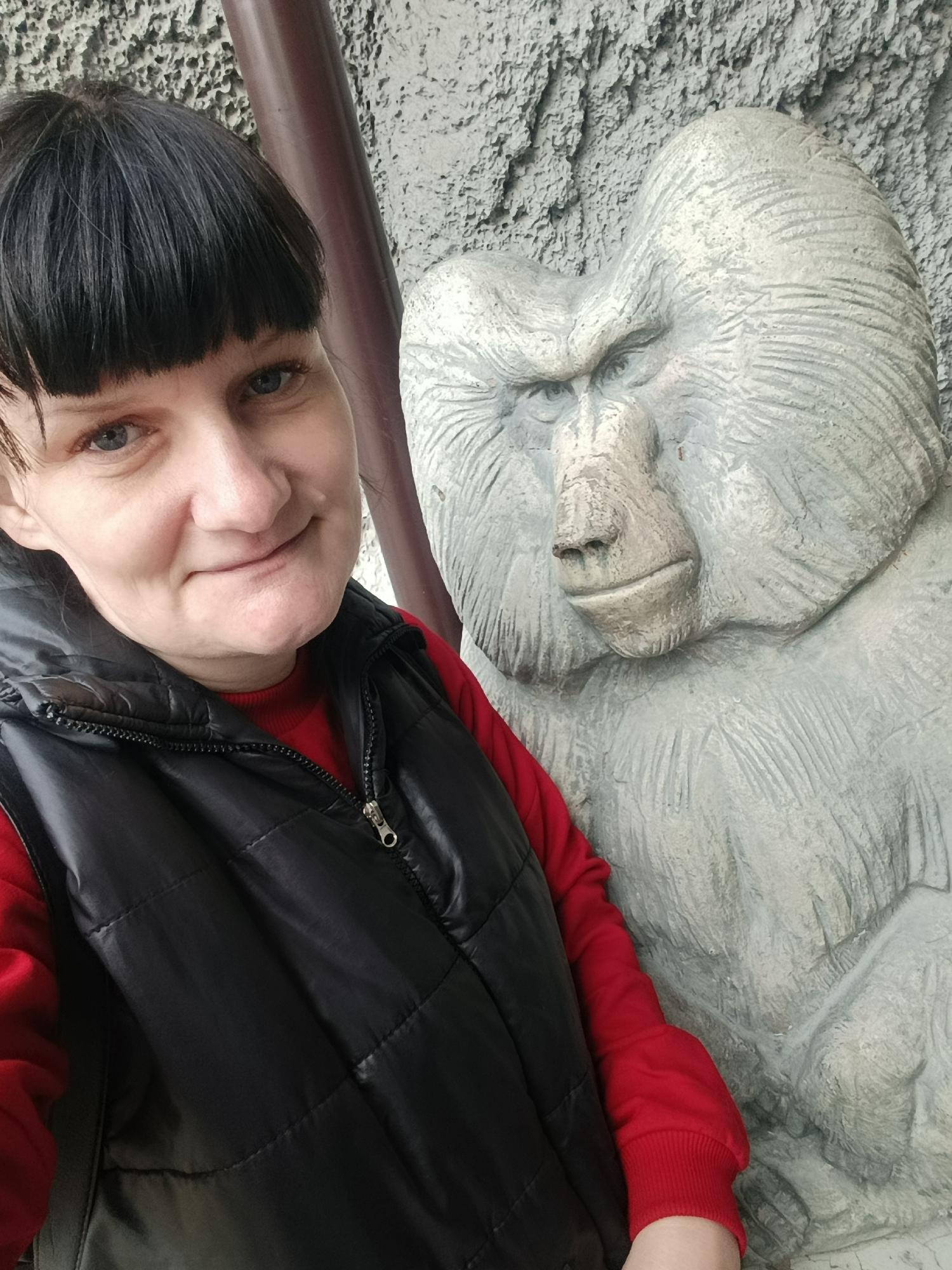 The image depicts a woman posing for a selfie next to a stone sculpture of a baboon. She is wearing a red sweater and a black padded vest. The background shows a textured wall that appears to be part of a larger structure or enclosure. The location is in Izhevsk, Russia.