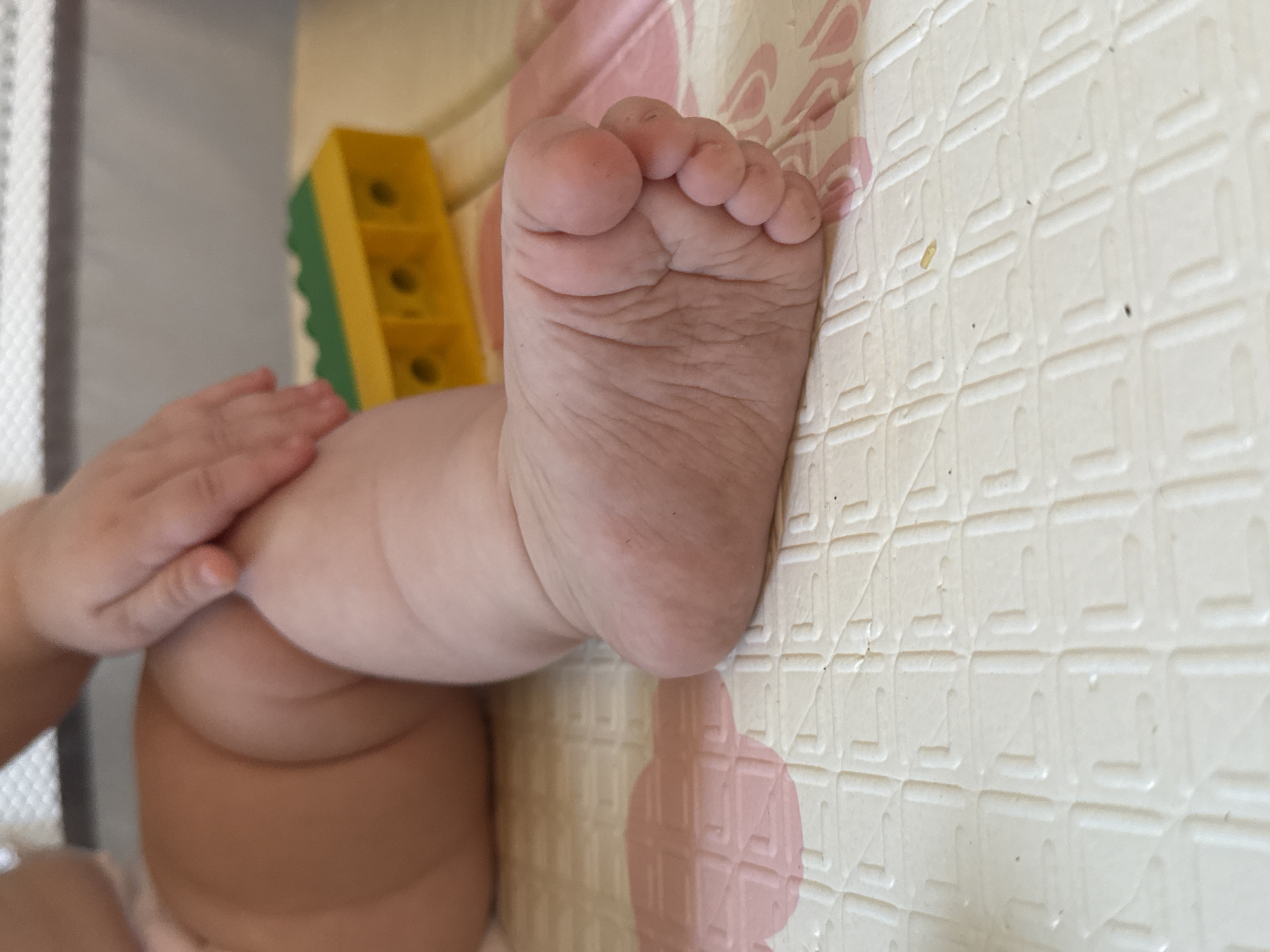 A baby's foot is prominently displayed against a patterned playmat. The foot is pink and fleshy, with distinct wrinkles and toes. To the left, the baby's hand and leg are partially visible. A yellow and green toy is also present near the leg. The playmat is light-colored with a grid-like pattern and pink accents. The location in the city of Rusalevka, Russia is not discernible from this image.