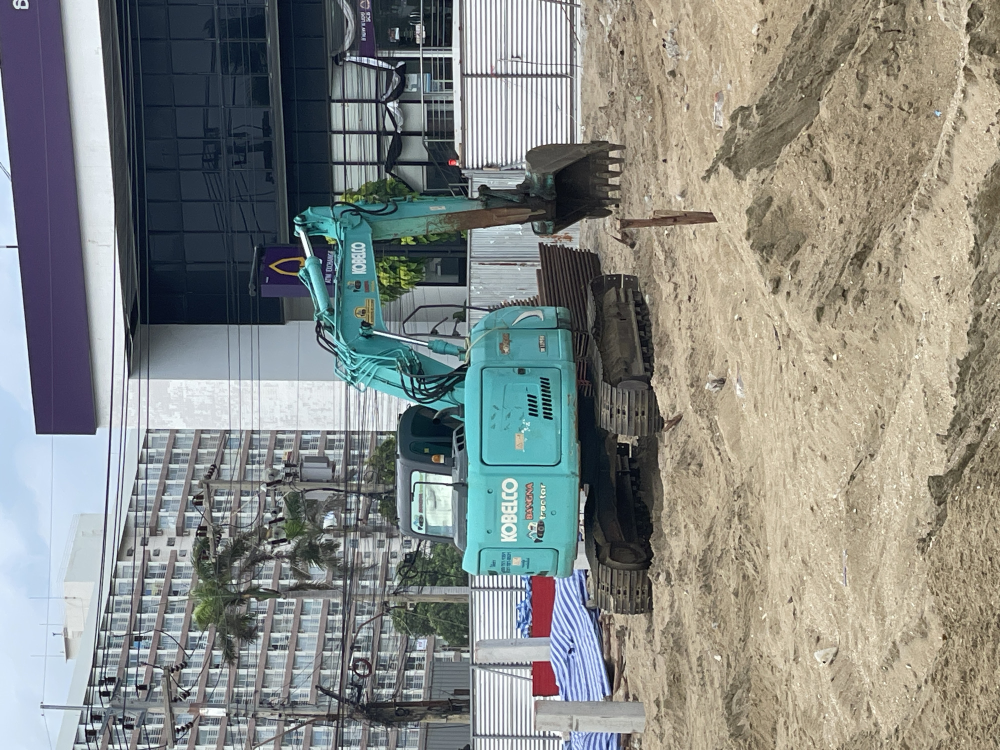 An outdoor, eye-level, medium shot captures a turquoise Kobelco excavator situated on a sandy construction site in Pattaya City, Thailand. The excavator is the central subject, its boom and bucket extended forward, positioned at a slight angle to the right. The machine bears the "KOBELCO" logo and the text "BANGNA CONTRACTOR".
In the background, a modern building with a dark glass facade and a purple awning is visible, with the words "ATM EXCHANGE" and a purple logo suggesting a financial institution. To the left of this building, a tall, multi-story residential building with numerous balconies and windows is seen, along with an intricate network of electrical poles and wires. Palm trees are interspersed between the buildings and utility poles.
The foreground and midground consist of uneven, sandy terrain, indicative of an active construction zone. Piles of sand are present, along with some construction materials like blue and white striped tarpaulins and metal poles. A few white concrete posts stand upright in the sand. The sky above is overcast with a light gray hue. There are no people visible in the image.