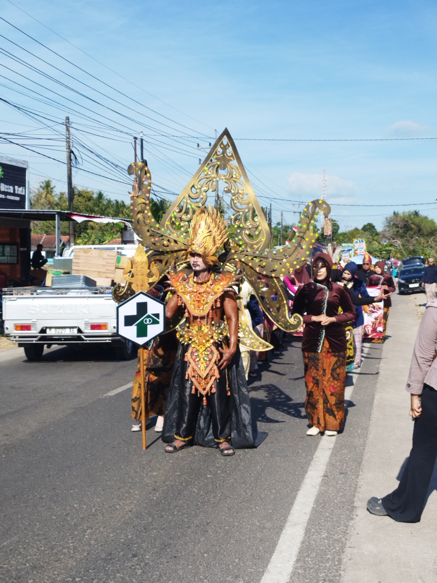 In the city of Mandala, Indonesia, a parade is in progress under a clear blue sky. The foreground is dominated by a person in an elaborate costume, featuring a golden headdress, ornate jewelry, and a flowing black skirt. They hold a staff with a hexagonal sign displaying a green cross and two intertwined rings. To the left, a white Suzuki Carry pickup truck is parked on the side of the road, its bed filled with boxes. Behind the main figure, a procession of people in traditional Indonesian attire walks along the asphalt road. Some individuals wear headscarves and long patterned skirts, while others are in darker, more formal clothing. In the background, trees and buildings are visible, along with overhead power lines. The overall scene depicts a cultural event or festival taking place on a sunny day.