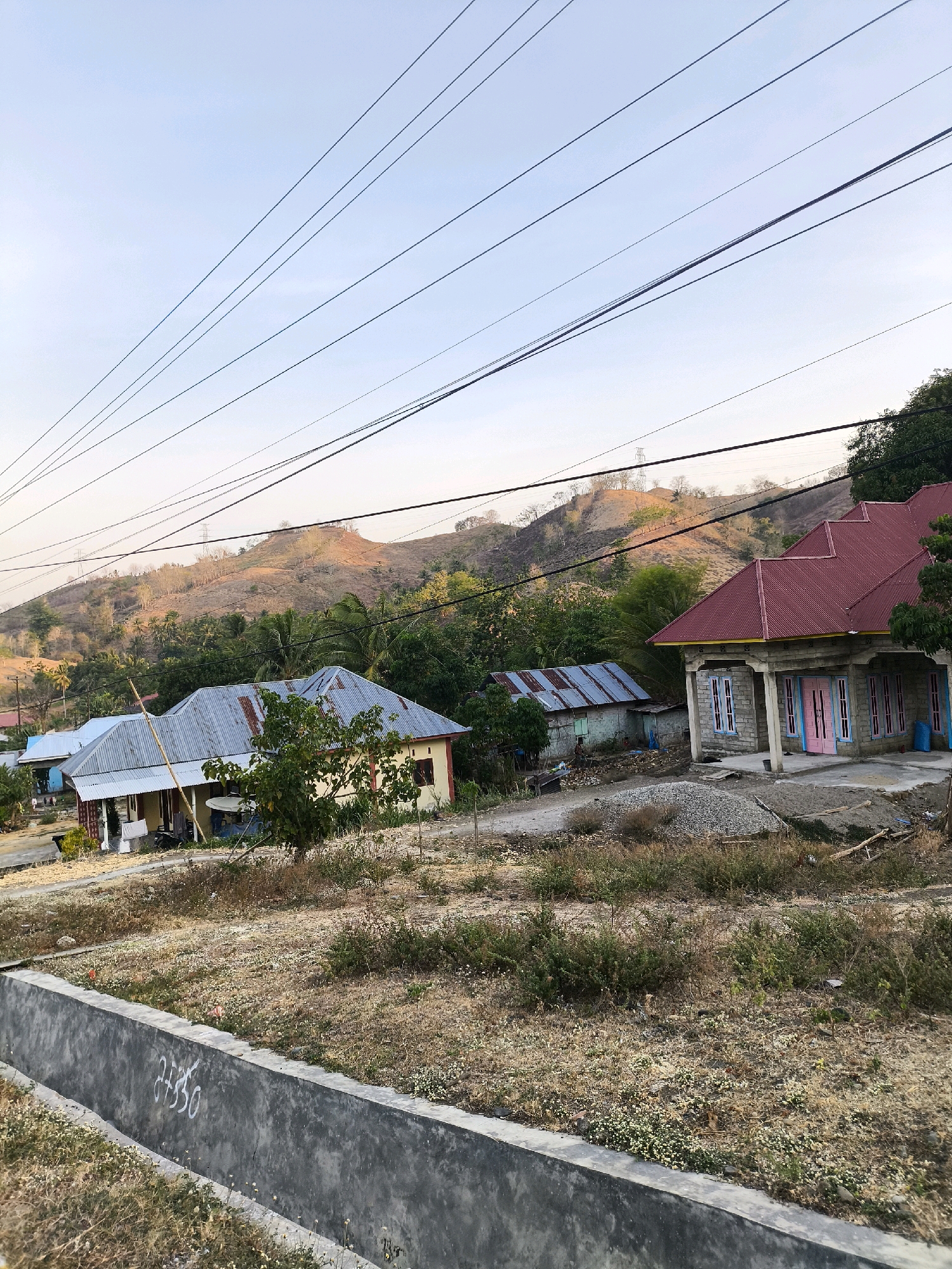 In the image, a rural Indonesian landscape is depicted under a pale sky. Multiple power lines crisscross the frame from left to right, overhead. In the middle ground, two houses are visible. The house on the left has a light-colored facade and a gray corrugated metal roof, with a porch supported by columns. To its right, a second house is more prominent, constructed with stone-like blocks and featuring a vibrant red corrugated metal roof. This house has several windows with light blue frames and pink doors. Adjacent to this house, a smaller structure with a gray roof is partially visible. The foreground features a low, dark gray concrete barrier with white markings, possibly a house number or identifier, written on it. Beyond the houses, a dry, hilly terrain is covered with sparse vegetation and scattered trees, with some patches of darker green foliage indicating denser tree cover. The overall impression is one of a developing rural area, possibly in a tropical or subtropical climate, given the vegetation and the style of the houses.