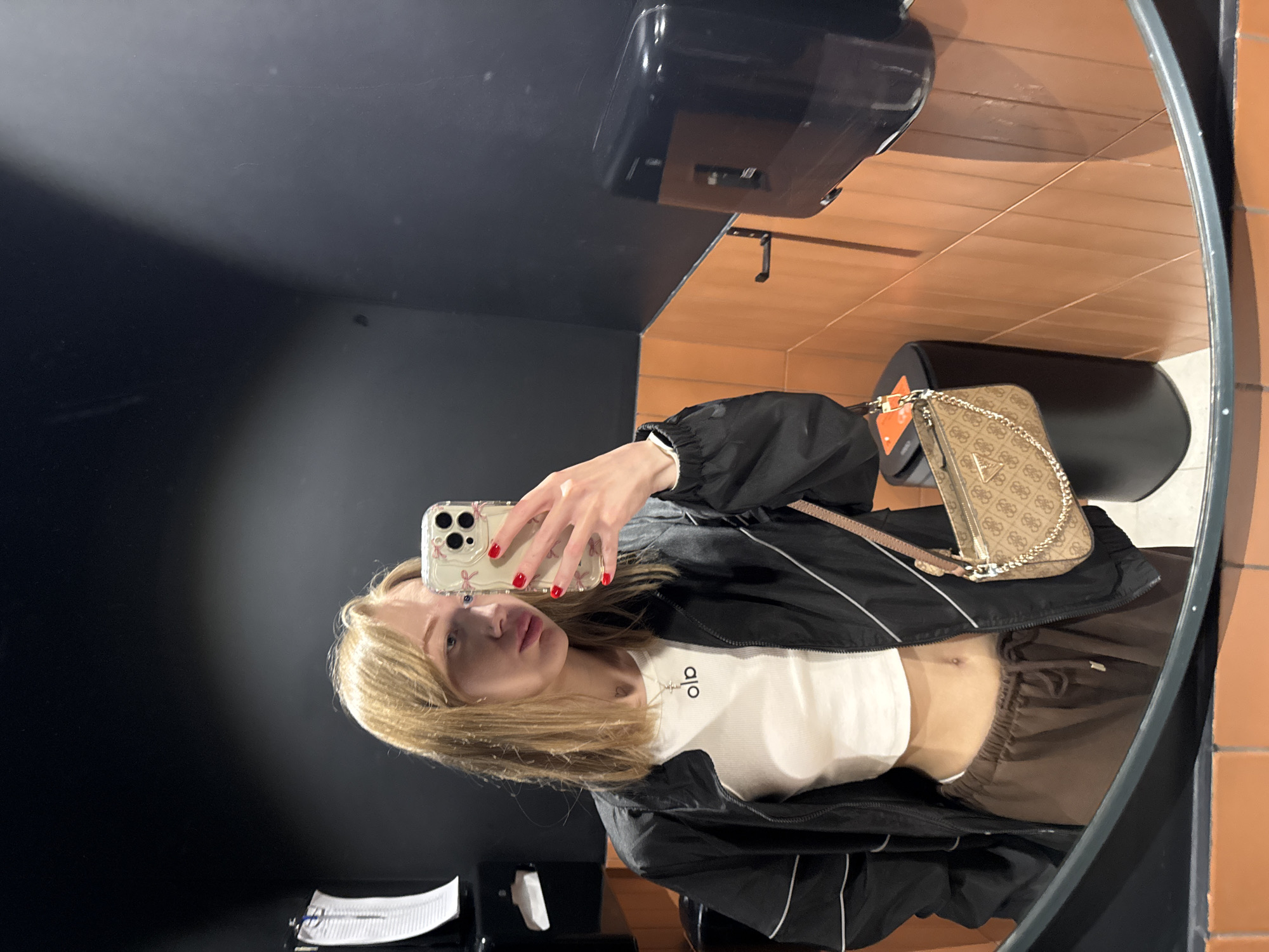 The image depicts a young woman taking a selfie in a restroom. She is wearing a white crop top with the word "alo" written in small letters, a black jacket with white accents, and brown sweatpants. Her blonde hair is shoulder-length, and she has red nail polish. She is holding an iPhone with a decorative case. Beside her, reflected in the mirror, is a beige patterned purse with a gold chain. The restroom has dark walls, a black automatic paper towel dispenser, and orange-tiled walls in the mirror's reflection. A black toilet is also visible in the reflection. There is a white tissue box and other small items on the counter under the mirror. The location within the city of Ovoshchnoy, Russia, cannot be confirmed from the image alone.