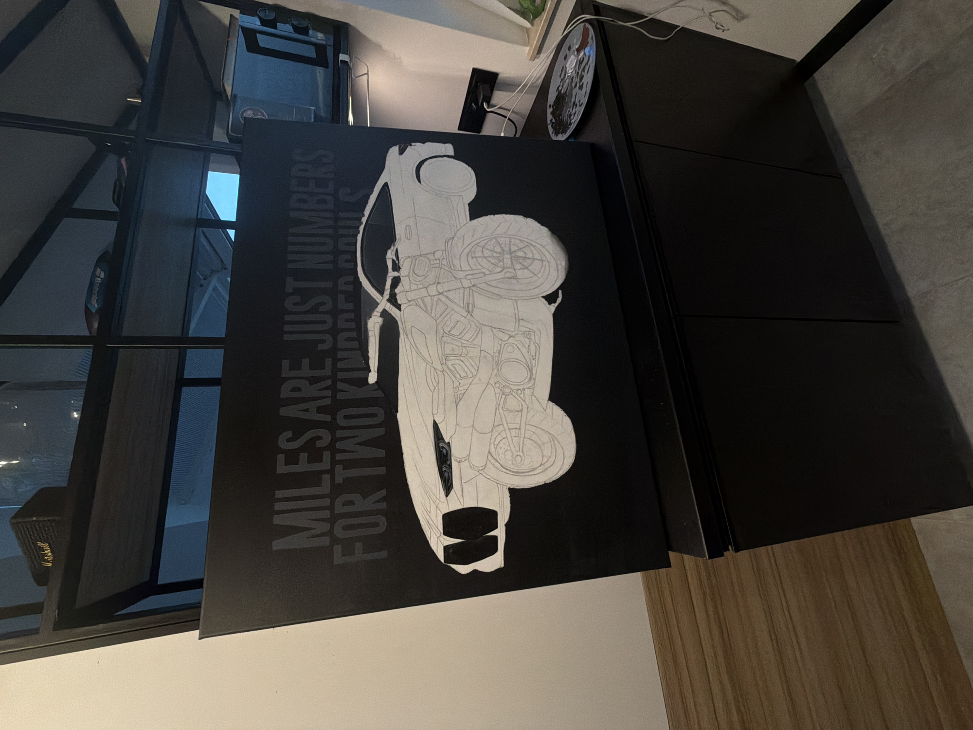 Here is a detailed description of the image:
The image shows a black canvas with white graphic of a motorcycle leaning against a dark-colored cabinet or shelving unit. The canvas bears the inscription "MILES ARE JUST NUMBERS FOR TWO KINDRED SOULS" in white text, positioned vertically alongside the motorcycle graphic. The motorcycle is rendered in an outline style, with key components like the tires, frame, and body visible.
The background features an indoor setting, suggesting a room in a house or apartment. A corner of the room is visible, with the cabinet or shelving unit taking up a significant portion of the right side. A vertical metal structure, likely a part of a larger shelving system or architectural element, is situated on the left. Natural light filters through what appears to be a window, casting a soft glow on the objects in the scene.
Several items are scattered around, including a clock and electronics that add to the domestic ambiance. The floor is tiled, offering a neutral, clean surface that contrasts with the darker furniture and canvas. The photograph appears to be taken at eye-level, providing a natural perspective of the objects and layout of the room.