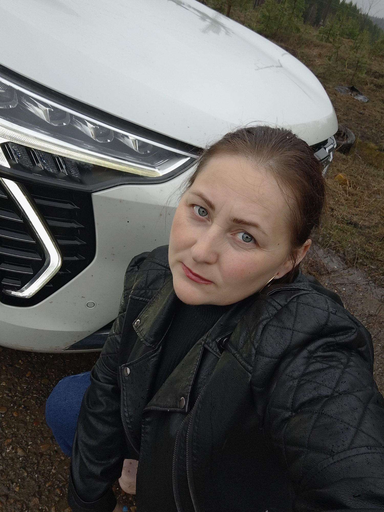 A woman is captured in a selfie-style shot, crouching or kneeling beside the front of a white car. She looks directly at the camera with a neutral to slightly serious expression, her blue eyes prominent. Her brown hair is pulled back. She is wearing a black quilted leather jacket, which has visible water droplets, suggesting rain or damp conditions. Underneath, she wears a black top, and blue jeans are visible on her lower body. Small gold hoop earrings adorn her ears.
The white car, likely an SUV or crossover, takes up a significant portion of the left and upper frame. Its modern LED headlights are illuminated, and part of its distinctive black grille with chrome accents is visible. The car's hood and bumper appear somewhat dirty with specks of mud, consistent with the outdoor, unpaved setting.
The background reveals a natural, somewhat unkempt environment. Bare or sparsely leaved trees are visible in the distance under an overcast sky, suggesting a cool or gloomy day. The ground around the car and the woman is muddy and wet, indicating recent rain or damp conditions. The overall scene suggests an outdoor location, possibly a rural track or unpaved area in Chaikovsky, Russia. There is no visible text.
