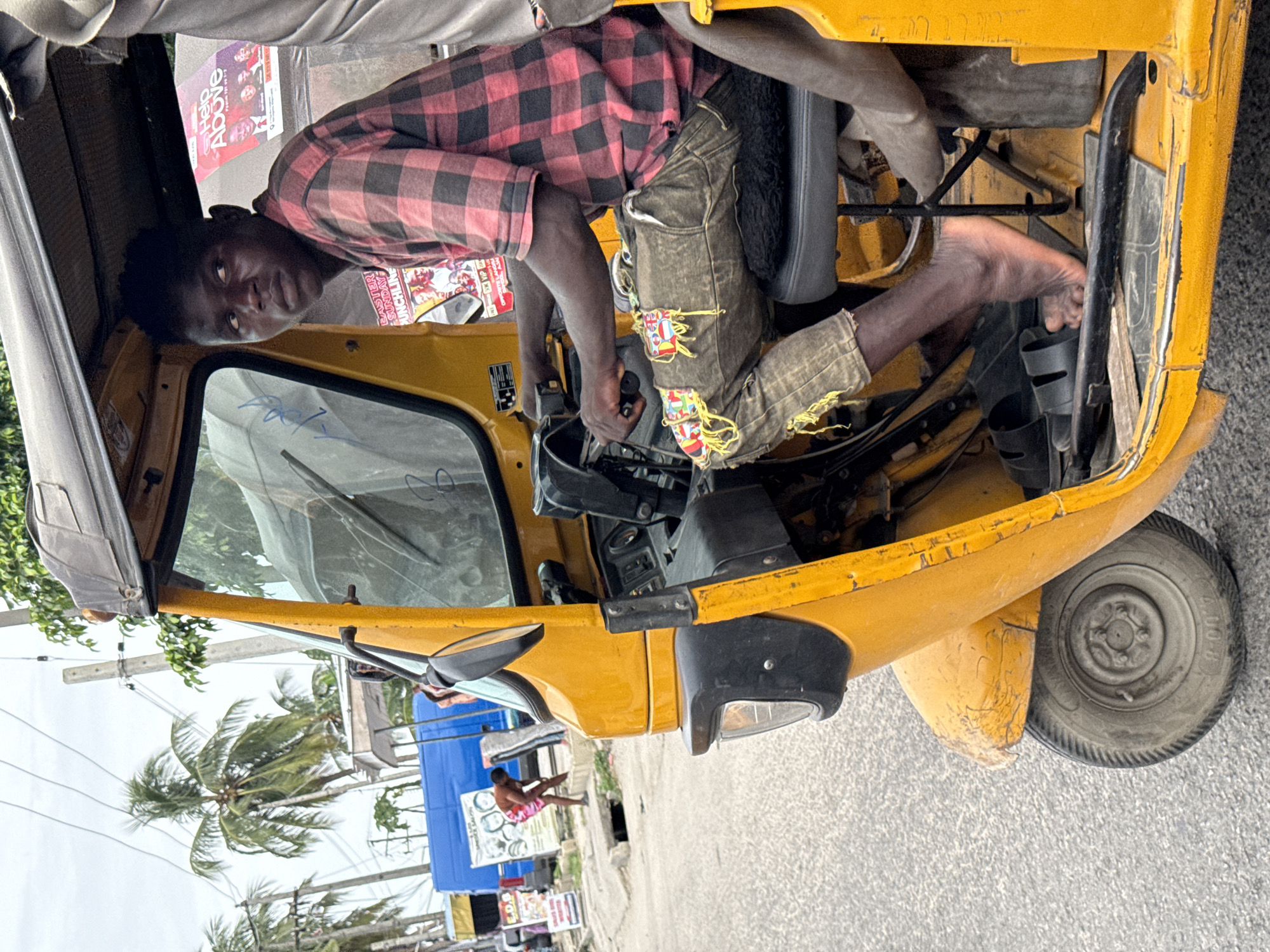 A young man in a red and black checkered shirt and distressed denim shorts sits in the driver's seat of a yellow auto rickshaw, commonly known as a keke napep, in Ado, Nigeria. His bare foot rests near the brake pedal, and he is looking at the viewer. Palm trees and a building with posters are visible in the background. The vehicle is on a street.