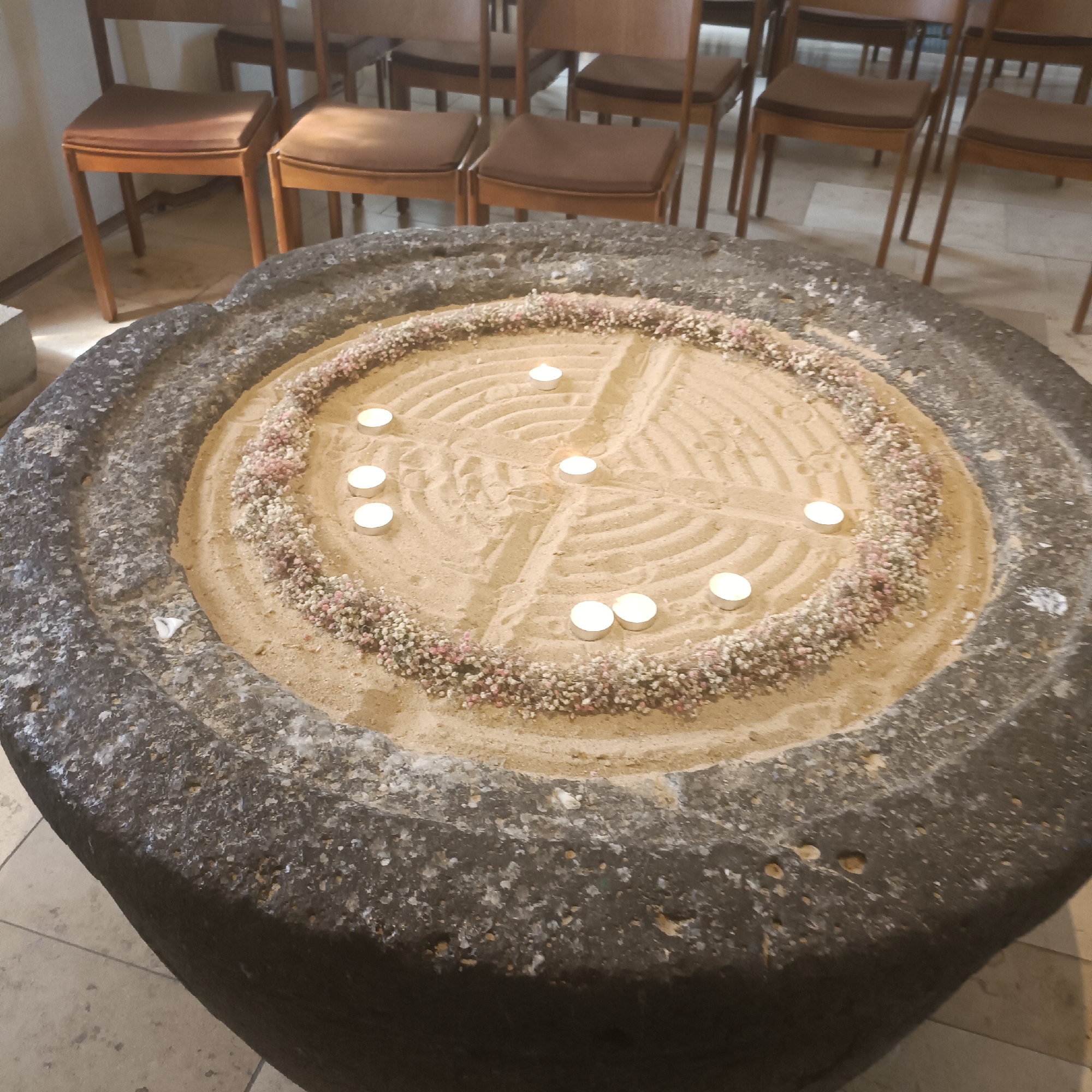 The image shows a circular stone basin filled with sand, decorated with a border of small white and pink flowers. Within the sand, a labyrinth pattern has been raked, and several small white tea lights are placed along the paths of the labyrinth, some of which are lit. In the background, wooden chairs with brown seats are visible, suggesting an indoor setting, possibly a church or a communal space. The overall impression is one of a contemplative or ceremonial arrangement.
This particular arrangement is located in the historic spa town of Bad Nauheim, Germany, specifically within the Trinkkuranlage. The Trinkkuranlage is a complex of buildings and gardens built in the Art Nouveau style, famous for its healing springs and elegant architecture. The stone basin with the labyrinth and candles is often used for spiritual reflection and meditation within the tranquil atmosphere of the Trinkkuranlage.