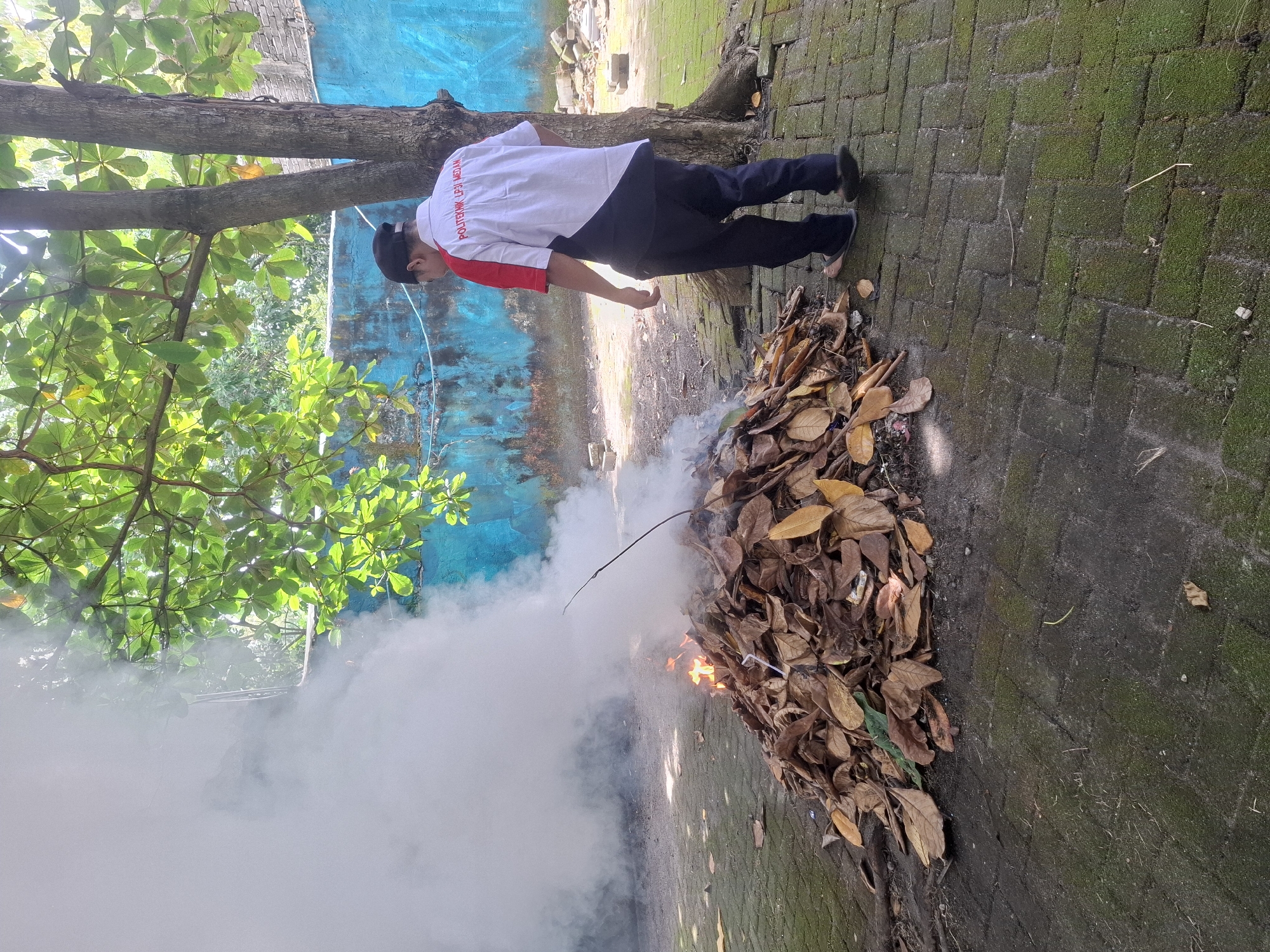 A person wearing a white and red shirt with "POLITEKNIK LP3I MEDAN" printed on the back, dark pants, and sandals stands near a pile of burning leaves, which is producing a large amount of smoke. The person is facing away from the camera and towards the fire. The scene appears to be outdoors in an area with a brick-paved ground covered in moss. A tree with green leaves is to the right of the person, and a blue wall is visible in the background. Scattered debris, including a white plastic straw, is present in and around the pile of leaves. The lighting suggests it is daytime, possibly morning or afternoon. The setting is likely within the grounds of Politeknik LP3I Medan in the city of Medan, Indonesia, given the text on the person's shirt.