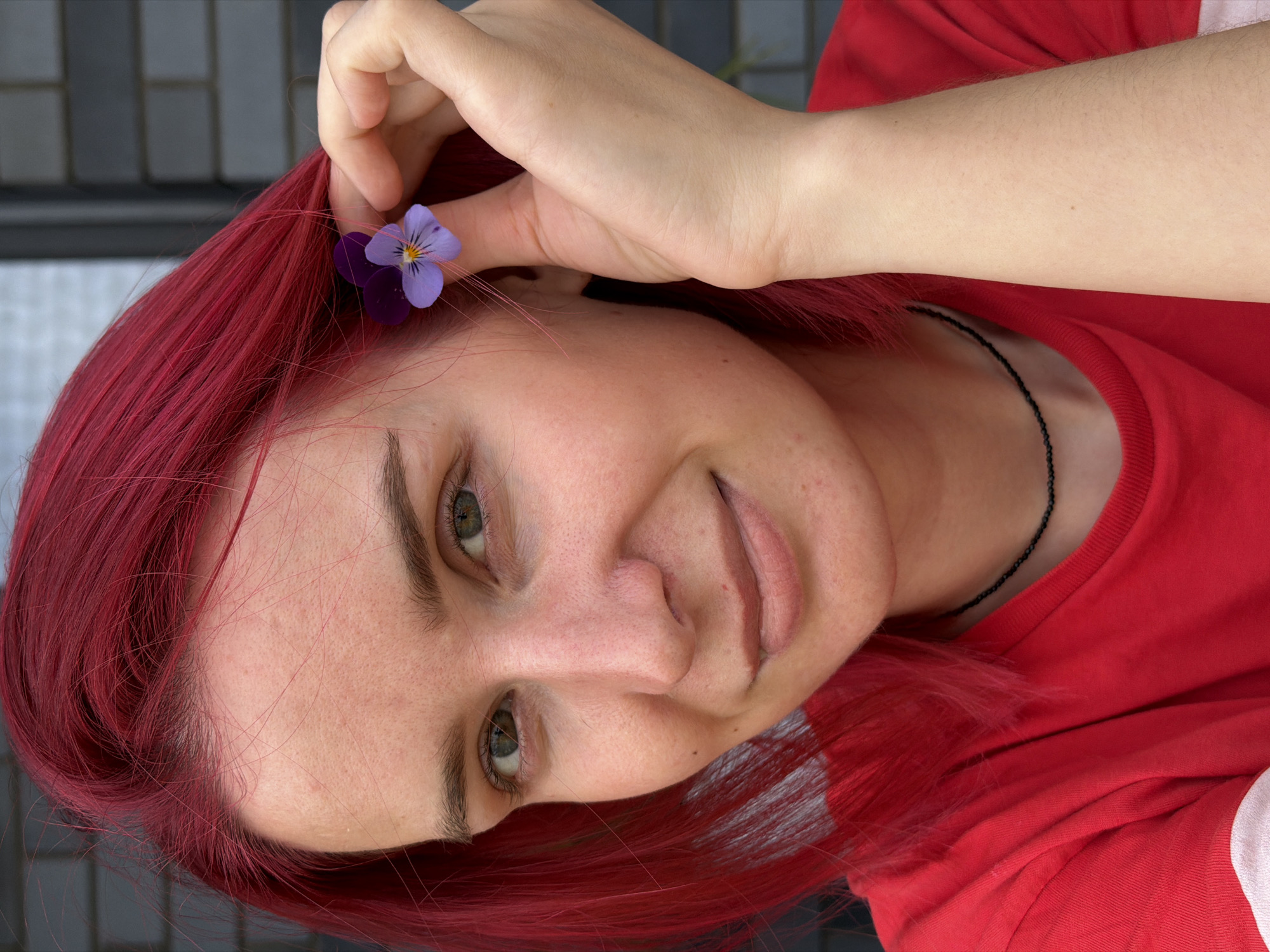 This is a close-up portrait of a young woman with bright red hair, who is holding a small purple and yellow pansy near her ear. She is wearing a red t-shirt and a thin black beaded necklace. Her skin is fair, with some visible pores and a few small blemishes. She has green eyes and is looking towards the camera with a slight smile. The background is out of focus and appears to be a brick wall. No specific location in the city of Chaikovsky, Russia, can be identified from this image.