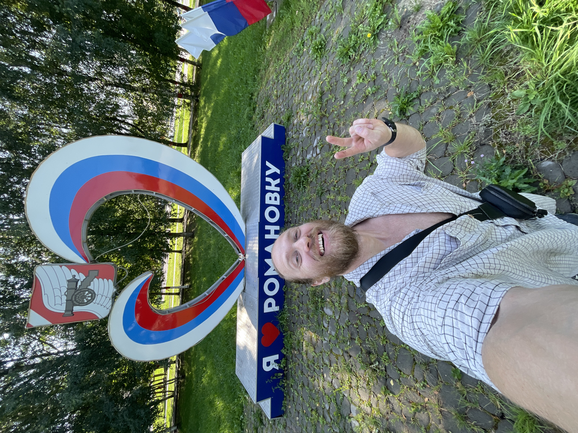 In the city of Romanovka, Russia, a man with a beard and wearing a checkered shirt is taking a selfie. He is standing in front of a large heart-shaped sculpture adorned with the colors of the Russian flag and a coat of arms featuring a cannon. The sculpture has text on a blue base that reads "Я ❤️ РОМАНОВКУ" (I love Romanovka). To the right of the sculpture, a Russian flag is visible. The man is smiling and making a peace sign with his fingers. The ground is paved with interlocking stones, and grass and trees are present in the background, suggesting an outdoor setting, possibly a park or public square.