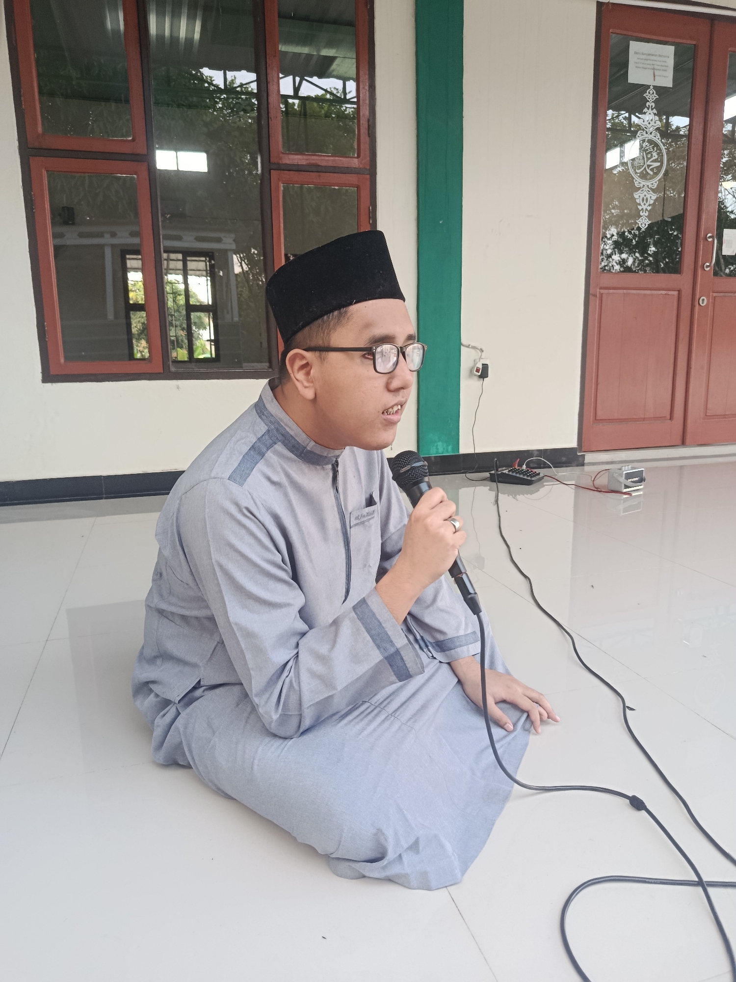 An Indonesian male, wearing a light gray Muslim robe and a black peci, sits on a white tiled floor in front of a building with large windows and double wooden doors. He is holding a black microphone and appears to be speaking. A black cable runs from the microphone towards the right side of the image. In the background, there is a small black audio mixer and a silver rectangular device connected by cables. The building's facade is white with a green vertical stripe. The windows reflect trees and sky. The scene is likely outdoors or in an open-air covered area. The location is in the city of Swarga Bara, Indonesia, possibly at a mosque or community center.