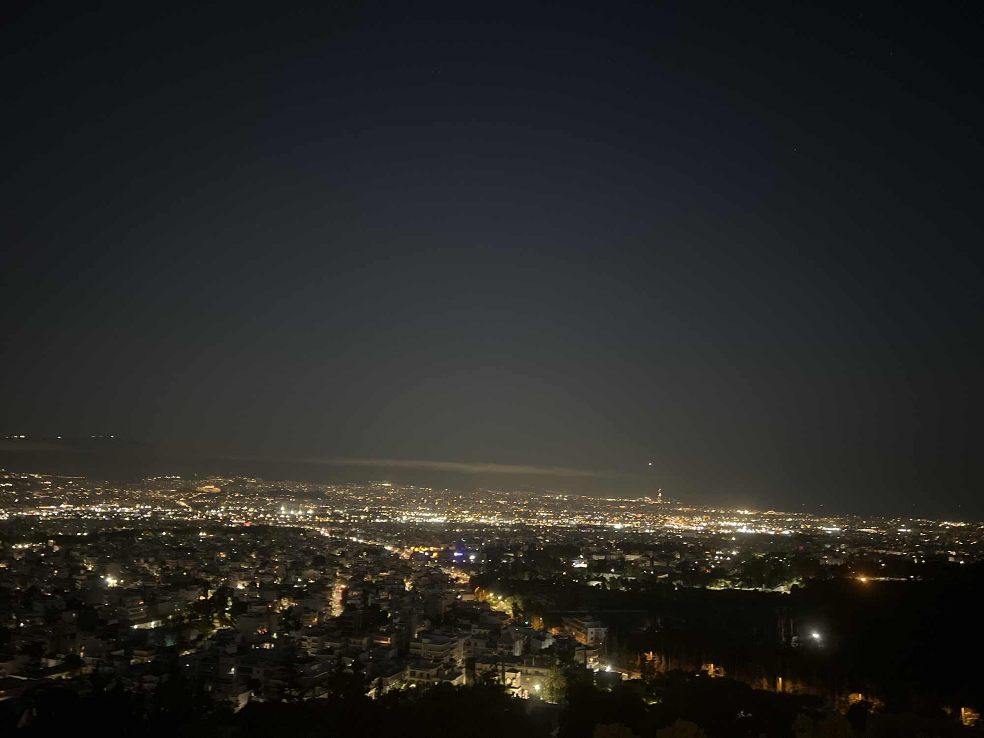 The image is a nighttime aerial view of a city, likely Athens, Greece, taken from a high vantage point. The foreground is dominated by dark silhouettes of trees and possibly some buildings on a hillside. Below, the city stretches out, a vast expanse of twinkling lights representing buildings, streets, and illuminated landmarks. The sky above is a dark, deep blue, with a few faint stars visible. In the distance, faint atmospheric haze can be seen, obscuring some of the city lights. The overall impression is one of a sprawling urban landscape at night, with the lights creating a warm glow against the dark sky. The location is identified as Chaidari, Greece, a municipality in the northwestern part of the Athens metropolitan area.