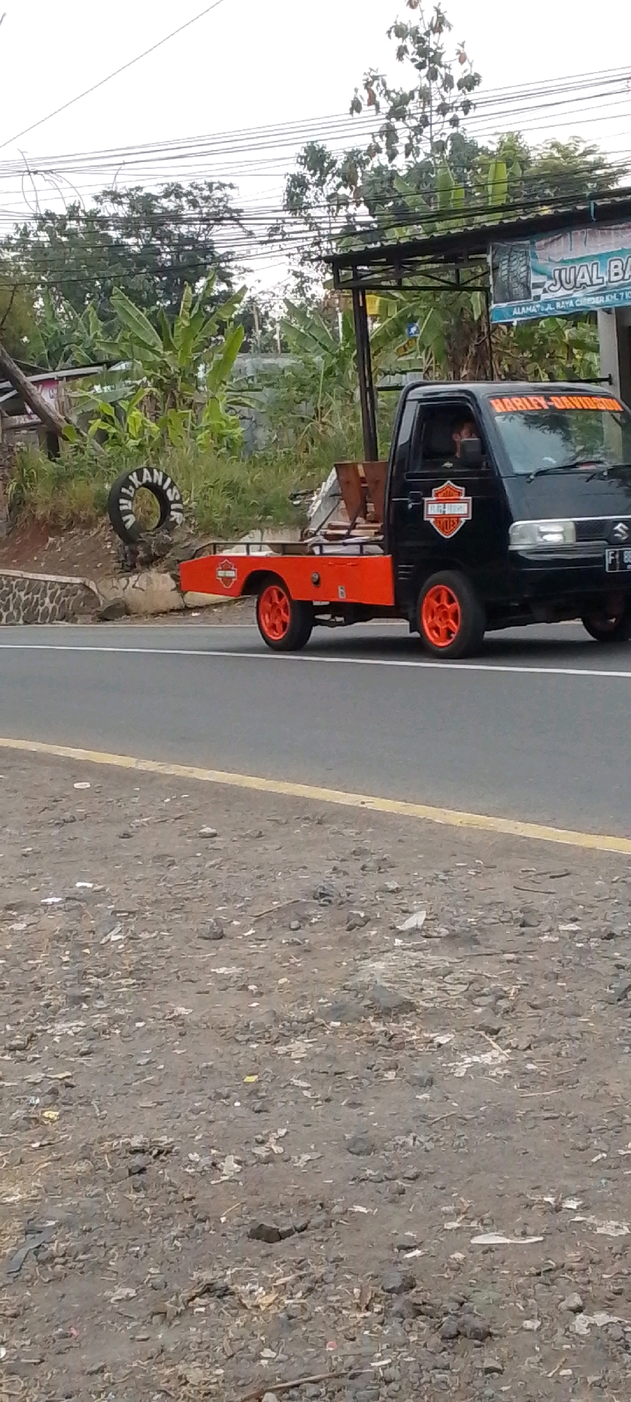 A black pickup truck with an orange flatbed, modified for hauling, is driving on a road in Cilaku, Indonesia. The truck has "HARLEY-DAVIDSON" decals on the windshield and a Harley-Davidson logo on its side. A person is visible in the driver's seat. The truck is adorned with multiple Harley-Davidson logos. The flatbed has some wooden materials loaded on it. In the background, lush green vegetation, including banana trees, is visible. A sign with text in Indonesian that translates to "JU AL BA" (likely part of "JUAL BARANG," meaning "FOR SALE") and an address is mounted on a structure to the right of the road. A discarded tire with text on it is positioned on the embankment to the left of the truck. The road is asphalt with a yellow line marking its edge. The surrounding environment appears rural or suburban, with power lines overhead. The ground next to the road is a rough, dirt-covered surface with scattered rocks and debris.