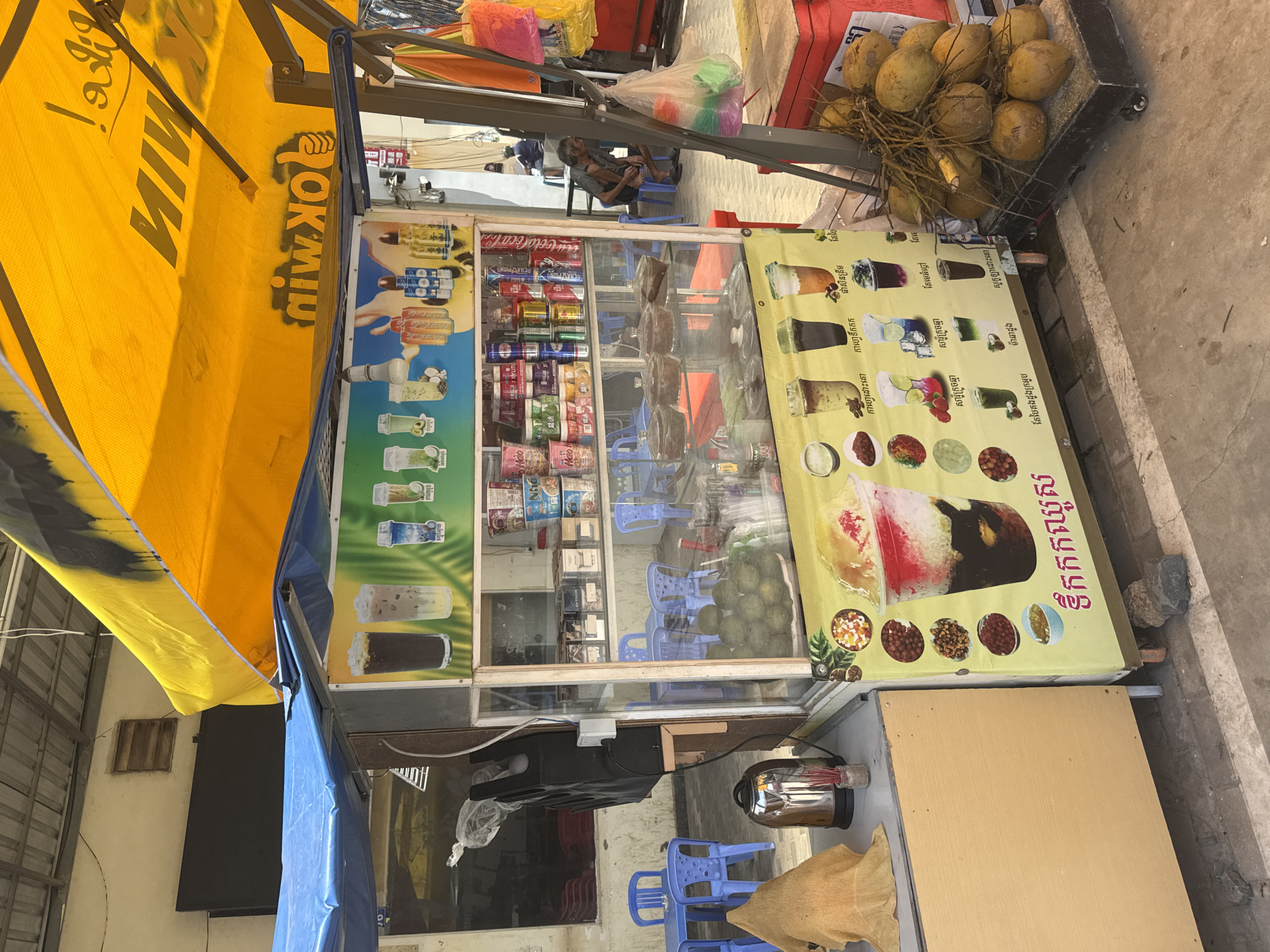 This is a street scene in Bavet Quarter, Cambodia, featuring a food and drink stall. The stall has a yellow canopy with black lettering that reads "OK MIN". The stall displays a variety of beverages in bottles and cans, as well as colorful illustrations of drinks on its front panel. There are also fruits, possibly coconuts, stacked to the right of the stall. In the background, a man is seated on a blue chair outside a building, and other blue tables and chairs are visible, suggesting an outdoor seating area. The ground is paved and appears to be a sidewalk or street. The overall impression is of a bustling marketplace or roadside establishment.