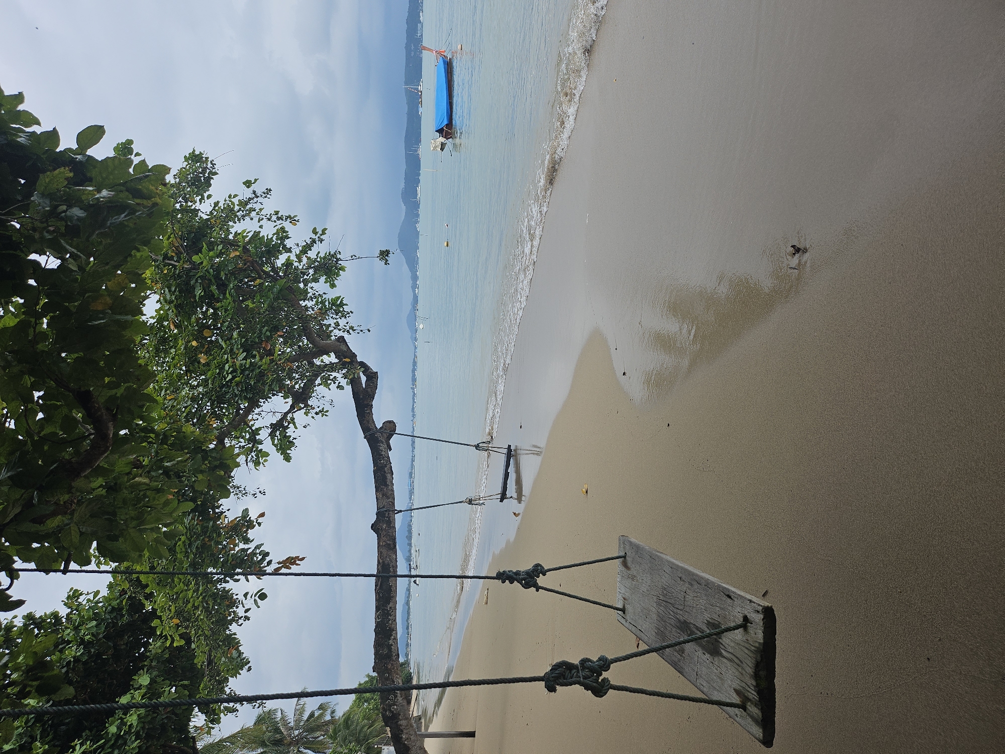 A serene, overcast day at a beach in Rawai, Thailand, featuring two wooden swings suspended from a large tree branch overhanging the sand. In the foreground, the left swing is closer to the camera, its weathered wooden seat and thick rope visible. A second, identical swing is further to the right, hanging over the wet sand closer to the water. The beach itself is a strip of light brown sand, with the gentle tide washing ashore in small waves. The water is a muted blue, dotted with numerous boats in the distance, including a longtail boat covered with a blue tarp. Palm trees are visible to the far left, and a hazy mountain range can be seen on the horizon under the cloudy sky. The overall scene evokes a sense of peaceful isolation and natural beauty.