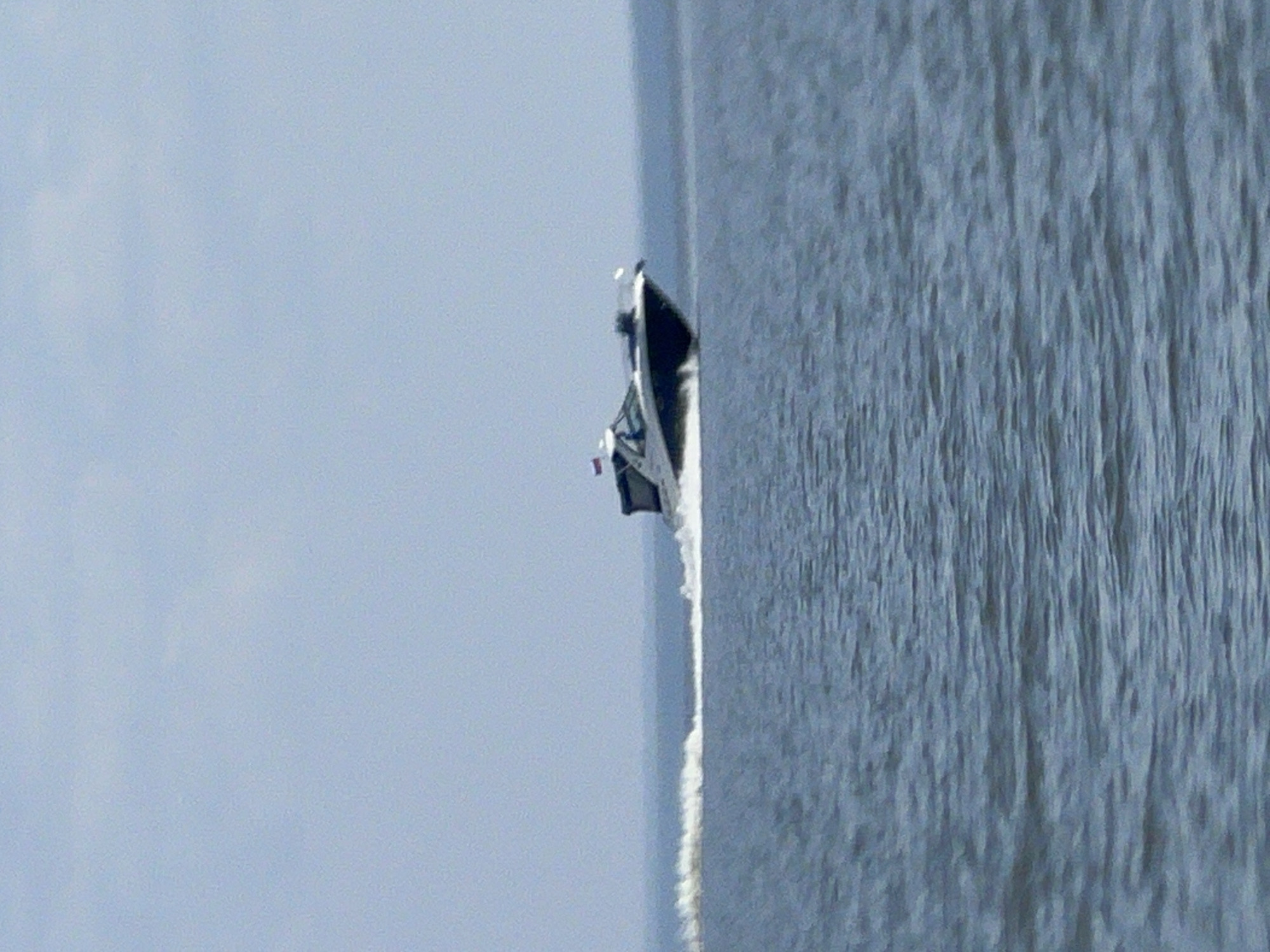 An outdoor daytime shot shows a motorboat speeding through calm water, leaving a white wake. The boat, which appears to be dark in color with a lighter cabin and windshield, has a Russian flag on its stern. The sky is a pale blue, and the water is a deep blue, with visible ripples. The scene is set on a large body of water, likely a lake or sea, under a clear sky. The location is not specifically identified as being in the city of Borovoe, Russia.