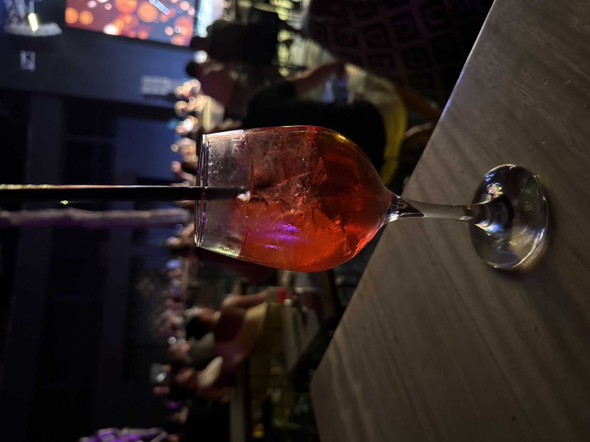A close-up, eye-level, medium shot captures a glass of red liquid with ice and a black straw, positioned on a wooden table in the foreground. The glass is in focus, showing reflections of pink and purple light from the ambient surroundings. The background is blurred and depicts a dimly lit, crowded indoor venue, likely a nightclub or bar. People are visible sitting at tables, with some standing. The overall atmosphere suggests a lively evening setting in Bodrum, Turkey, possibly at a venue along the coast, given the typical nightlife scene there.
