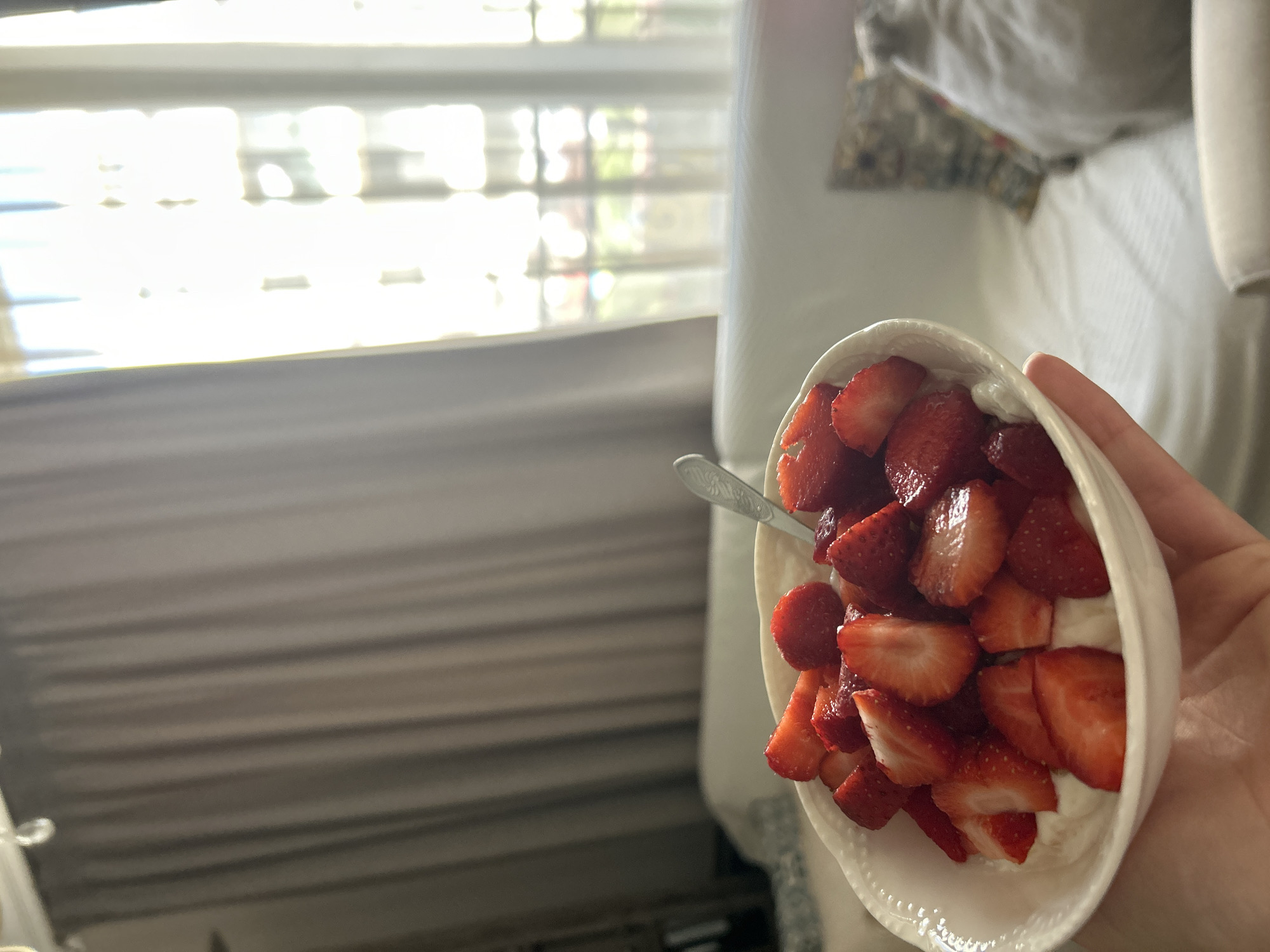 A hand holds a white, ornate bowl filled with sliced strawberries over a bed. The bowl is white with a raised, decorative pattern around the rim. The strawberries are bright red and appear fresh, sliced into pieces. A silver spoon with an ornate handle rests in the bowl, partially submerged in the strawberries. Behind the bowl, a plush white couch is visible, and to the left, a grey curtain is drawn. Sunlight streams through a window with sheer white curtains, creating a bright, hazy effect. The scene suggests a cozy indoor setting. The location is not discernible from the image.