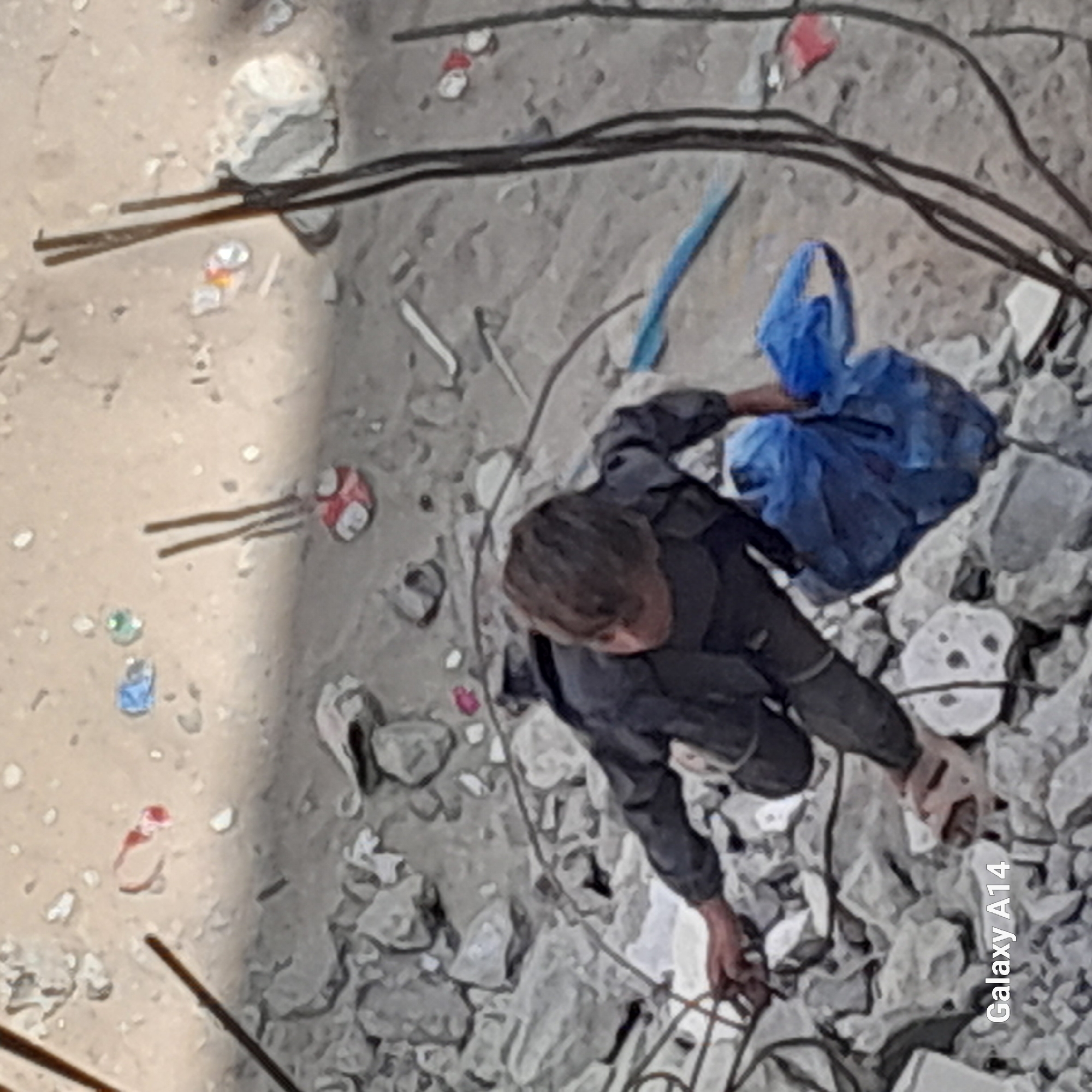 The image depicts a scene of urban devastation, potentially in the city of Unknown, Unknown. The ground is covered in rubble and debris. Amidst the ruins, a person wearing dark clothing is crouched down, sifting through the rubble with their hands. A blue bag is nearby. Electrical wires are visible amidst the debris. The overall scene suggests a disaster or conflict zone.