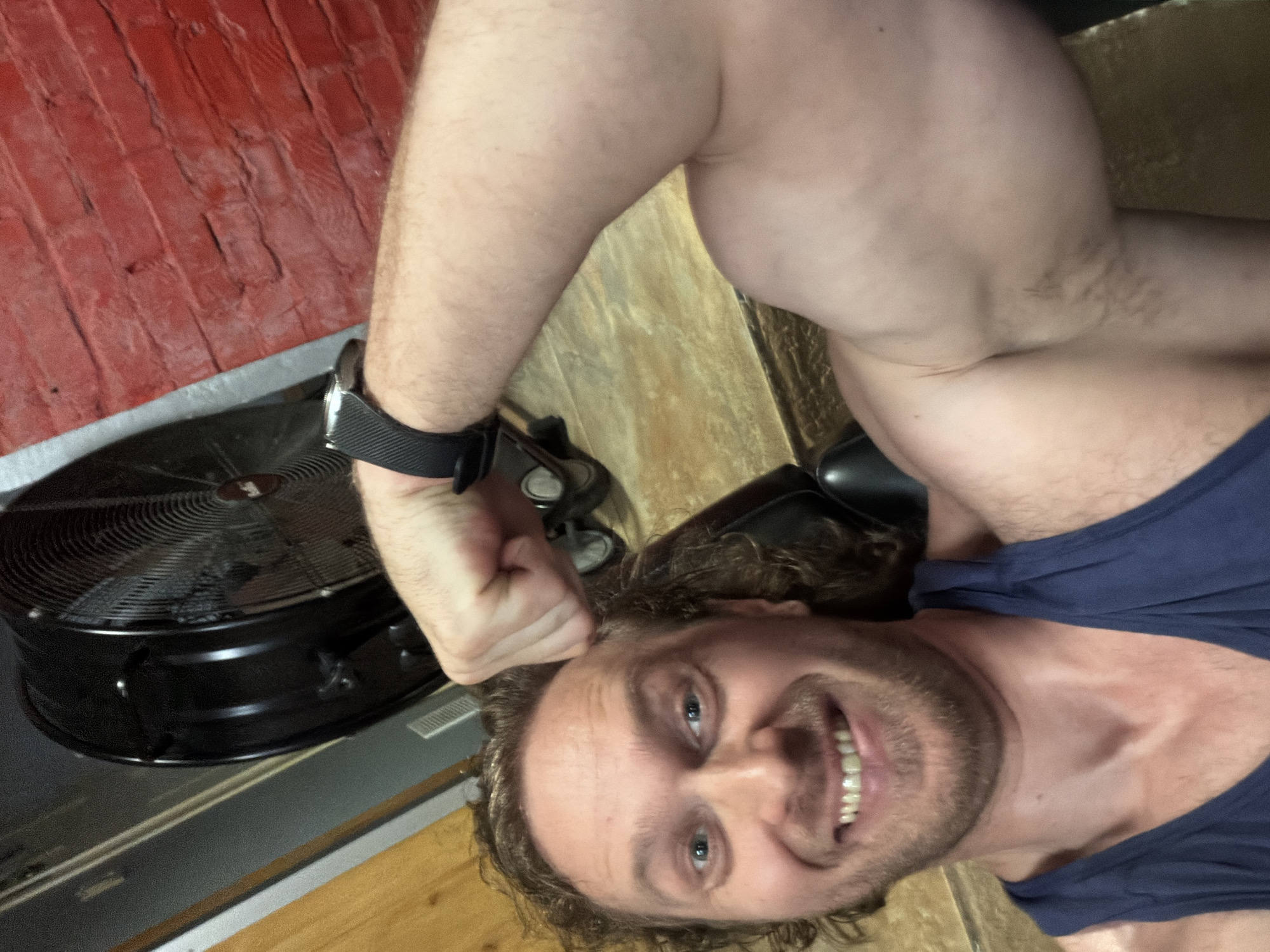 A man takes a selfie from a low-angle perspective, smiling broadly with white teeth, revealing light blue eyes and curly brown hair. He has stubble or a short beard and is wearing a dark blue tank top. His left arm is flexed, showcasing his bicep, with a black smartwatch on his wrist.
The background features a textured red brick wall on the upper left, possibly painted or stained. A light-colored wooden or laminate floor occupies the lower and right sections of the image. A black, circular floor fan with a metallic grate is visible on the left side of the frame, near the wall. A silver strip or molding separates the wall and floor. The setting appears to be indoors, possibly a personal workout area. The man's expression is cheerful and confident. No legible text is visible in the image.