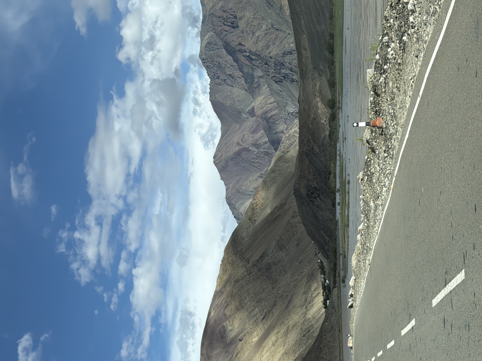 A paved road with white dashed and solid lines runs diagonally from the lower left to the mid-right of the frame. The road is bordered on the right by a rocky embankment. Beyond the embankment, a body of water, possibly a river, stretches across the midground. On the far bank of the water, a small cluster of buildings, likely a village, is visible at the base of a large, barren mountain. The mountain dominates the background, displaying layers of rock in various shades of brown and gray, with some areas cast in shadow. The sky above is a bright blue, dotted with scattered white clouds. A few small, reflective poles are visible along the embankment. The scene appears to be in Chumathang, India, a region known for its stark, mountainous landscapes and river valleys.