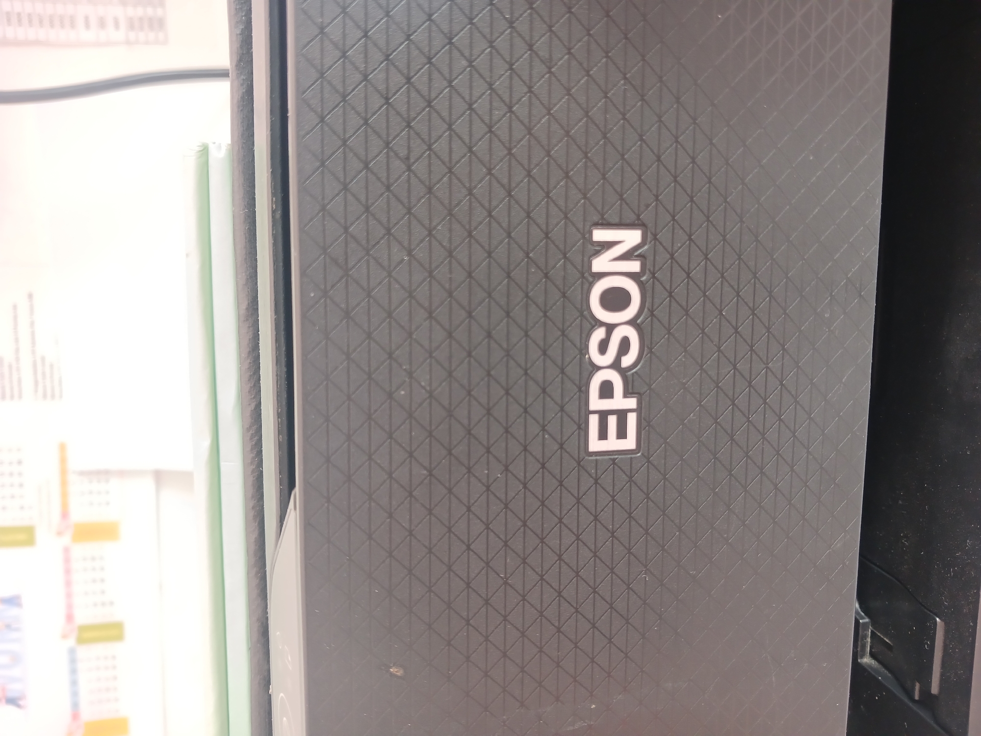 The image shows a close-up view of the top surface of a black Epson printer. The surface has a textured pattern of interlocking triangles. The word "EPSON" is printed in large, white, raised letters with a subtle pink outline, centered on the textured surface. The background is out of focus, but appears to be a wall with some papers or documents tacked up, possibly calendars or schedules, with some yellow and green accents. There is a hint of a window or bright light source in the upper portion of the image. The bottom of the image shows the edge of the printer and a black surface below it, possibly a desk or another part of the printer. The scene appears to be indoors, likely in an office or home setting. There are no people visible in the image. The image does not contain any information that would allow for the determination of its location within Watugede, Indonesia.