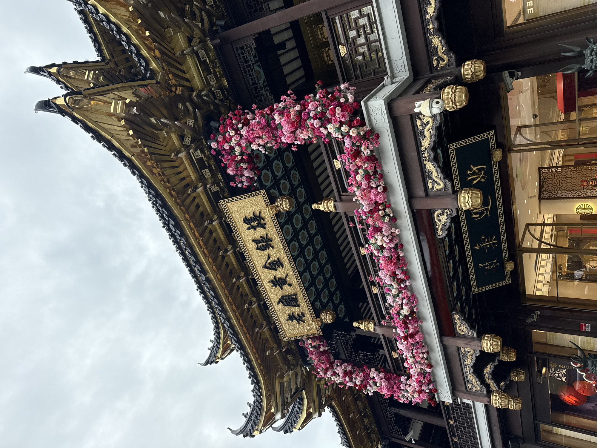 The image shows the exterior of a traditional Chinese building, likely a shop, in Shanghai, China. The building features ornate, tiered golden roofs with dark tiled eaves, a common architectural style in the region. The facade is adorned with intricate wooden carvings, many of which are gilded with gold, depicting dragons and other traditional motifs.
A prominent feature is a large floral arrangement composed of numerous pink and red flowers, which frames a balcony area. This floral display appears to be a festive decoration, possibly for a special occasion or holiday. Above the balcony, a golden plaque with Chinese calligraphy is mounted on the building. Below the balcony, another dark plaque with golden Chinese characters is visible.
The building's lower level has large glass windows and doors, revealing the interior which appears to be a brightly lit retail space. Security cameras are mounted on the exterior walls. The sky above is overcast and grey. The overall impression is one of traditional Chinese architecture embellished for celebration, located in a bustling urban setting. The specific location is likely within a historic or shopping district of Shanghai.