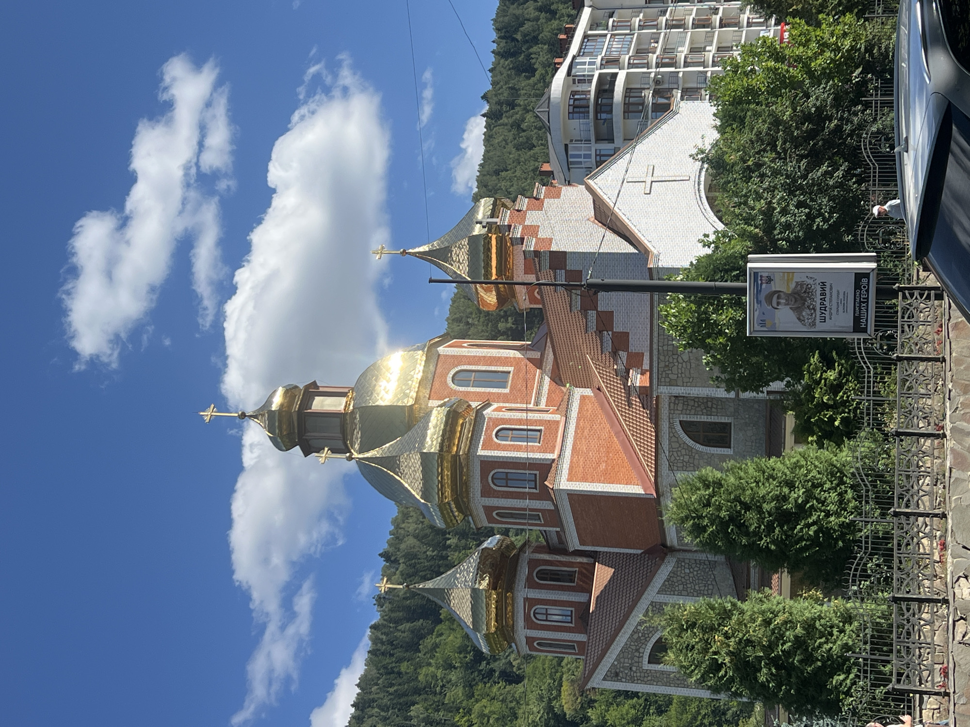 The image shows a prominent Ukrainian Orthodox Church with golden domes, situated in Yaremche, Ukraine. The church is constructed with red brick and stone accents. To the right of the church, a modern multi-story apartment building is visible. In the foreground, a black metal fence with decorative patterns runs along a stone wall. A billboard displaying a portrait of a Ukrainian soldier with text in Ukrainian is positioned near the fence. Several evergreen trees are planted in front of the church. The background features a lush, tree-covered hillside under a bright blue sky with scattered white clouds. A silver car is parked in the lower right corner of the frame.