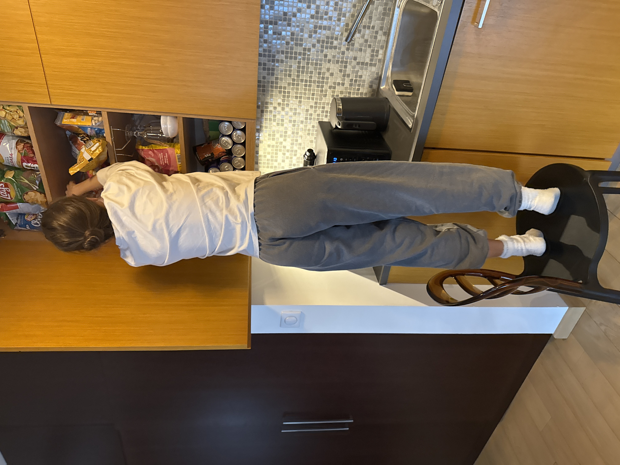 A person in a white shirt and gray pants reaches into a cupboard filled with food items, including chips and canned goods. They are standing in a kitchen next to a counter, sink, and a bar stool. The location is Warsaw, Poland, based on the mosaic tiles on the wall.