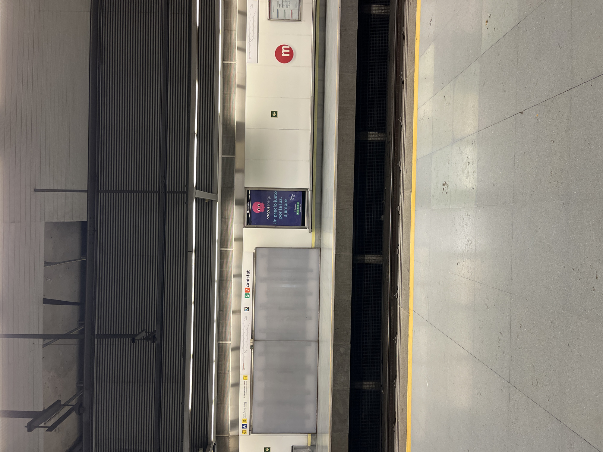 The image shows a view of a platform at the Amistat metro station in València, Spain. The platform is empty, with a yellow safety line marking the edge. Doors with "5 7 Amistat" are present, denoting the station and lines served. An advertisement for Octopus Energy is displayed. The station's logo, a red "m" within a circle, is visible. The platform floor is made of grey tiles. The ceiling has a ridged design with linear lighting fixtures.