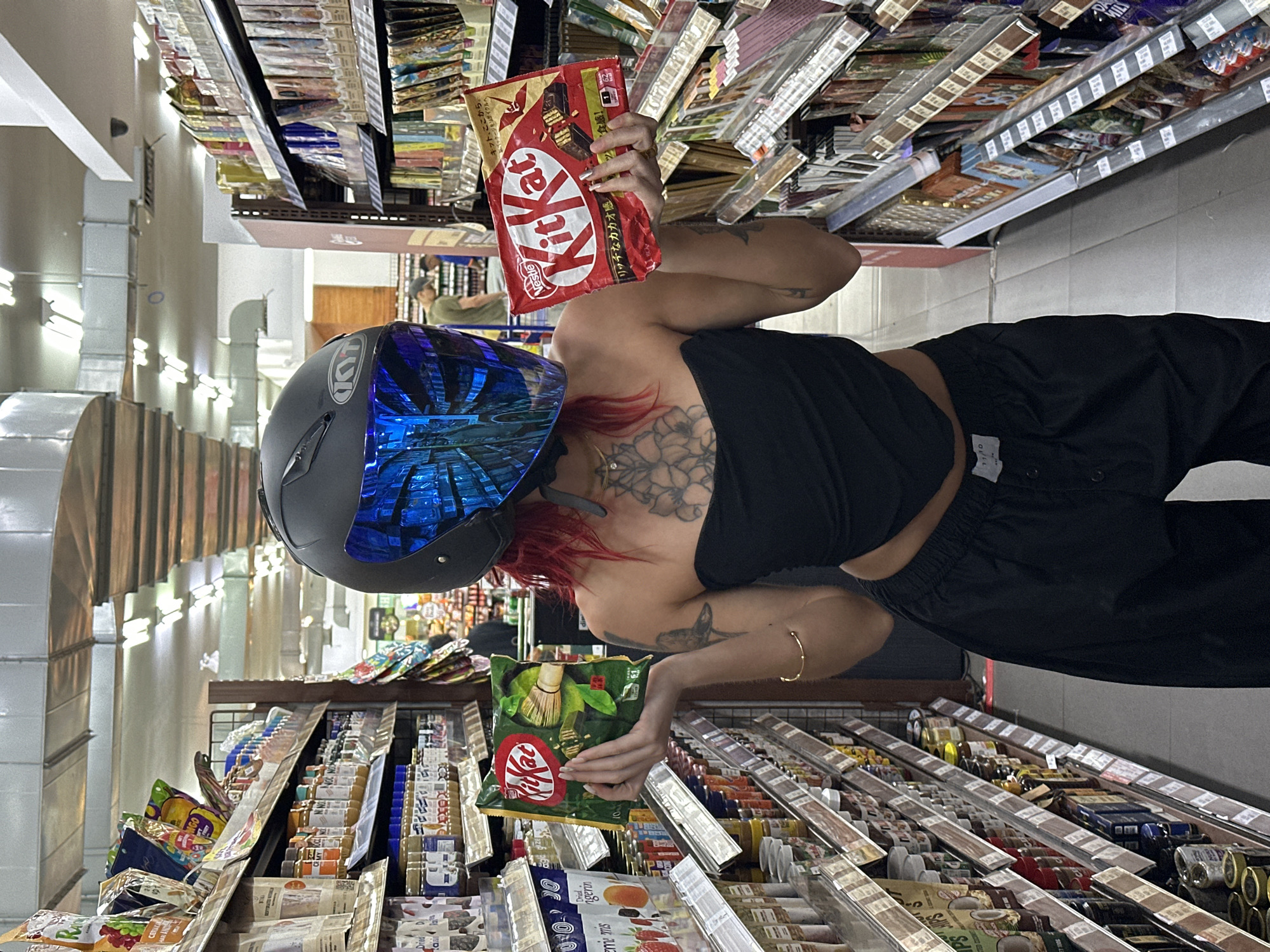 The image depicts an individual wearing a motorcycle helmet with a reflective blue visor, standing in a supermarket aisle, potentially in North Kuta, Indonesia. They are holding two different flavors of Kit Kat candy bars; one appears to be a Japanese import in red packaging, held above, and the other, a green tea flavor, held below. The individual is wearing a black top and dark pants. Their hair is dyed red, and they have several tattoos visible on their upper body. The supermarket aisle is stocked with various food products on shelves. The image orientation is rotated, making the supermarket aisles appear vertical and the person horizontal.