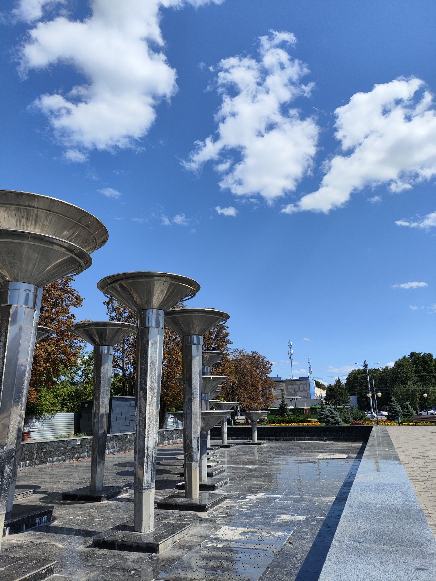 This is a vertical, eye-level, outdoor, daytime, full-shot photograph of a public fountain area in Kharkiv, Ukraine. The foreground features a series of large, metallic, cone-shaped structures, possibly lights or decorative elements of a dry fountain, arranged in a row. They are supported by metallic columns and concrete bases, all situated on a dark, tiled surface that appears wet in places. Behind these structures, there is a low, dark tiled wall. Further back, trees with autumnal foliage and green conifers are visible, as well as a grey corrugated metal fence. In the middle ground, there are buildings, including one with a large hexagonal pattern, and cell towers. A green bus, cars, and a park bench are also visible. The background consists of a bright blue sky with scattered white cumulus clouds. The overall impression is one of a well-maintained urban public space under a clear sky. The specific location in Kharkiv is believed to be Freedom Square (Ploshcha Svobody), a prominent central square known for its fountains and monuments.