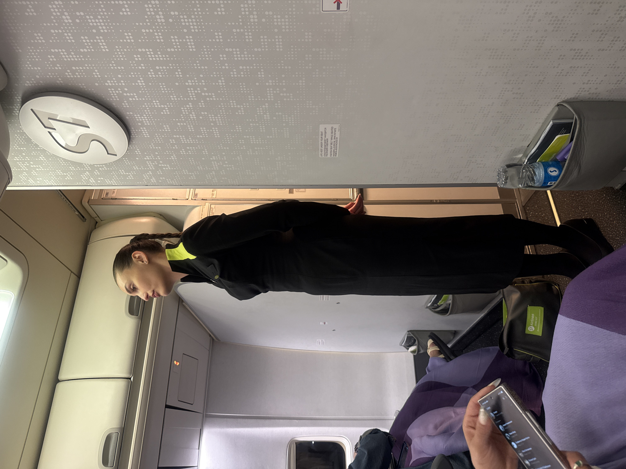 The image depicts the interior of an airplane cabin, specifically showing a flight attendant standing in the aisle. The flight attendant is wearing a black uniform with bright yellow accents on the collar and sleeves. She has her hair in a braid and is looking to her right. The cabin features overhead storage compartments, and the wall has a patterned texture with the "S7 Airlines" logo prominently displayed. Several passenger seats are visible, some with purple and blue seat covers. A passenger's hand holding a smartphone is in the foreground on the left. To the right of the flight attendant, a pocket is attached to the wall containing a water bottle, a glass, and reading materials. The scene is indoors, on an aircraft, and there is no indication of the specific location within the city of Artyom, Russia, beyond it being within an S7 Airlines plane.