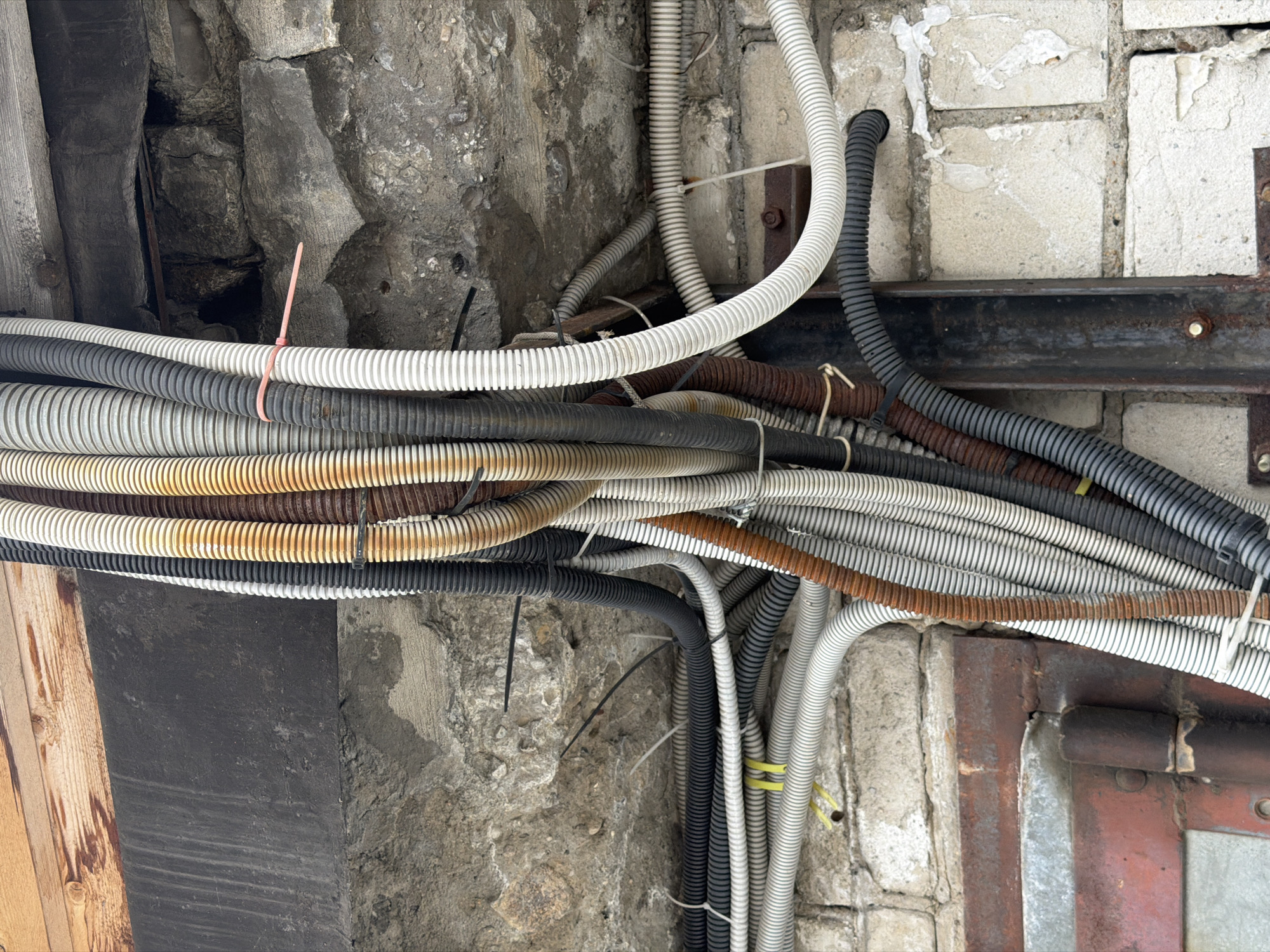 The image depicts a chaotic arrangement of corrugated electrical conduits running along a wall, presumably inside a building. The conduits are of varying colors: white, gray, black, and brown. They are bundled together with plastic zip ties. The wall is constructed of concrete and brick, with visible signs of wear and weathering. A rusted metal support structure is present near the brick section of the wall. A wooden surface is present on the left side of the image. There is no evidence of Birobidzhan, Russia in this image. The location cannot be determined from the image. No people, subjects, objects, actions, or interactions can be determined from this image other than the presence of the above materials.