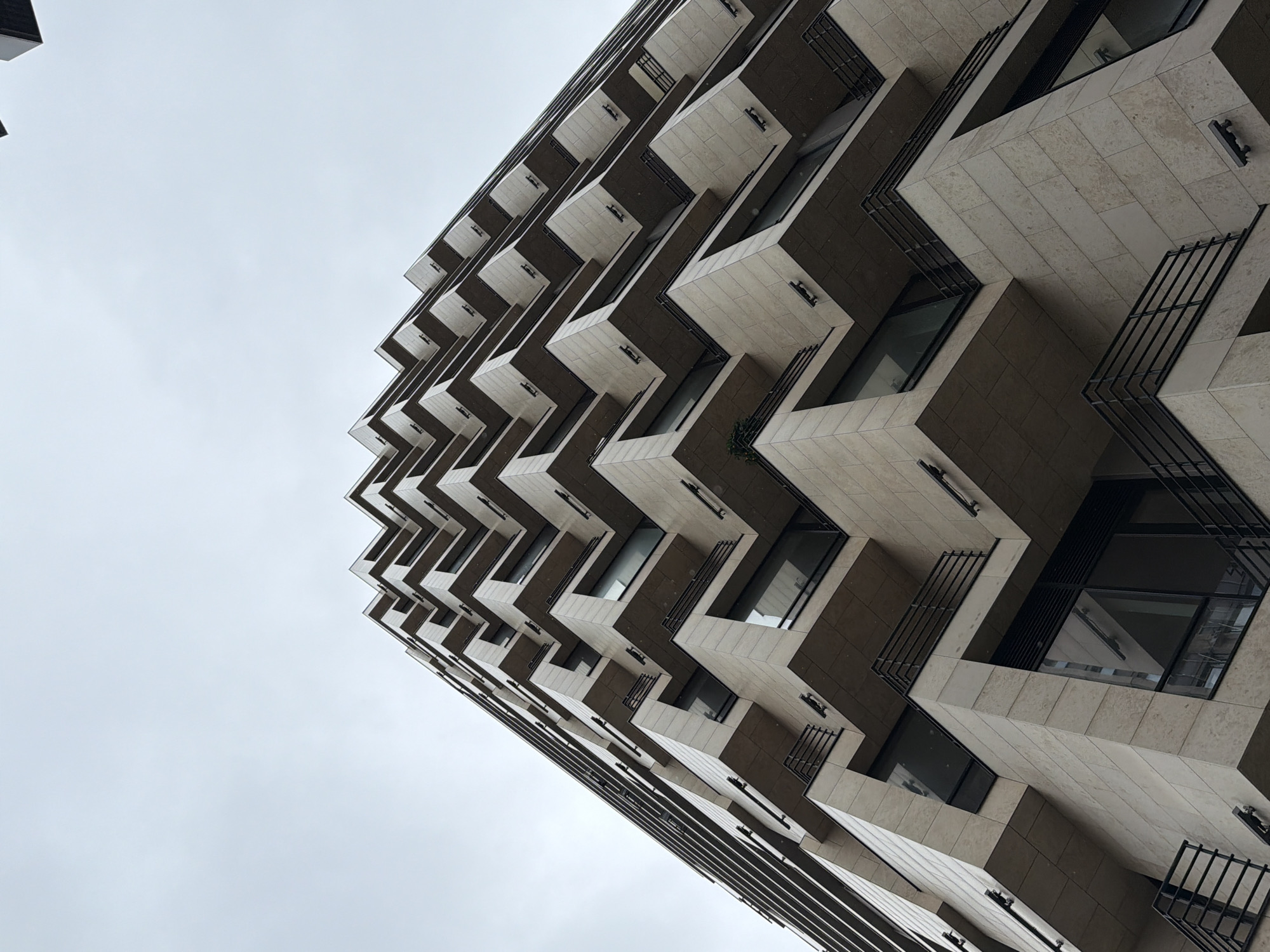 A low-angle shot of a modern building with a unique facade design. The building appears to be made of light-colored stone or concrete. The facade is composed of angular, protruding sections that create a zigzag pattern. Each section has a window and a small balcony with black metal railings. The sky is overcast. The building is in Moscow, Russia.