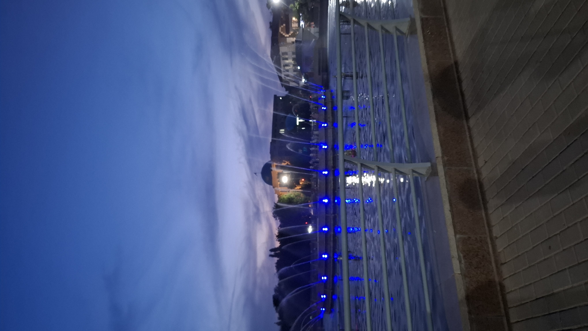 The image depicts a fountain display at dusk in Shymkent, Kazakhstan. The fountain features numerous jets of water, illuminated by bright blue lights, creating a captivating visual effect. The water arcs and sprays upwards, reflecting the blue lights and the twilight sky.
In the background, a building with a dome-shaped roof is visible, along with trees and other structures, suggesting an urban park or public space. The time of day is clearly evening, with the sky displaying a deep blue hue, transitioning into a softer twilight tone towards the horizon. Wispy clouds are present in the sky, adding texture to the dusky atmosphere.
The foreground shows a metal railing structure adjacent to the fountain, with a brick wall to its right. The water in the fountain area is calm, but the illuminated jets create a dynamic and lively scene. The overall mood is serene and magical, enhanced by the interplay of light and water. No people are clearly visible in the frame, and there is no discernible text. The scene is likely a public attraction, designed to be enjoyed in the evening.