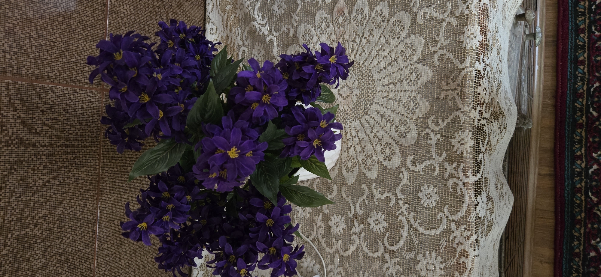 An artificial bouquet of dark purple flowers with yellow centers is arranged in a white pot. The pot sits on a table covered with a light-colored lace tablecloth that has an intricate floral pattern. Behind the bouquet, a wall is covered in a textured, brownish material, possibly wallpaper or fabric. The scene appears to be indoors, in a domestic setting, potentially a living room or dining area. The lighting is moderate, casting soft shadows on the objects. No people are present in the image. The location of Tel, Azerbaijan is not discernible from the provided image.