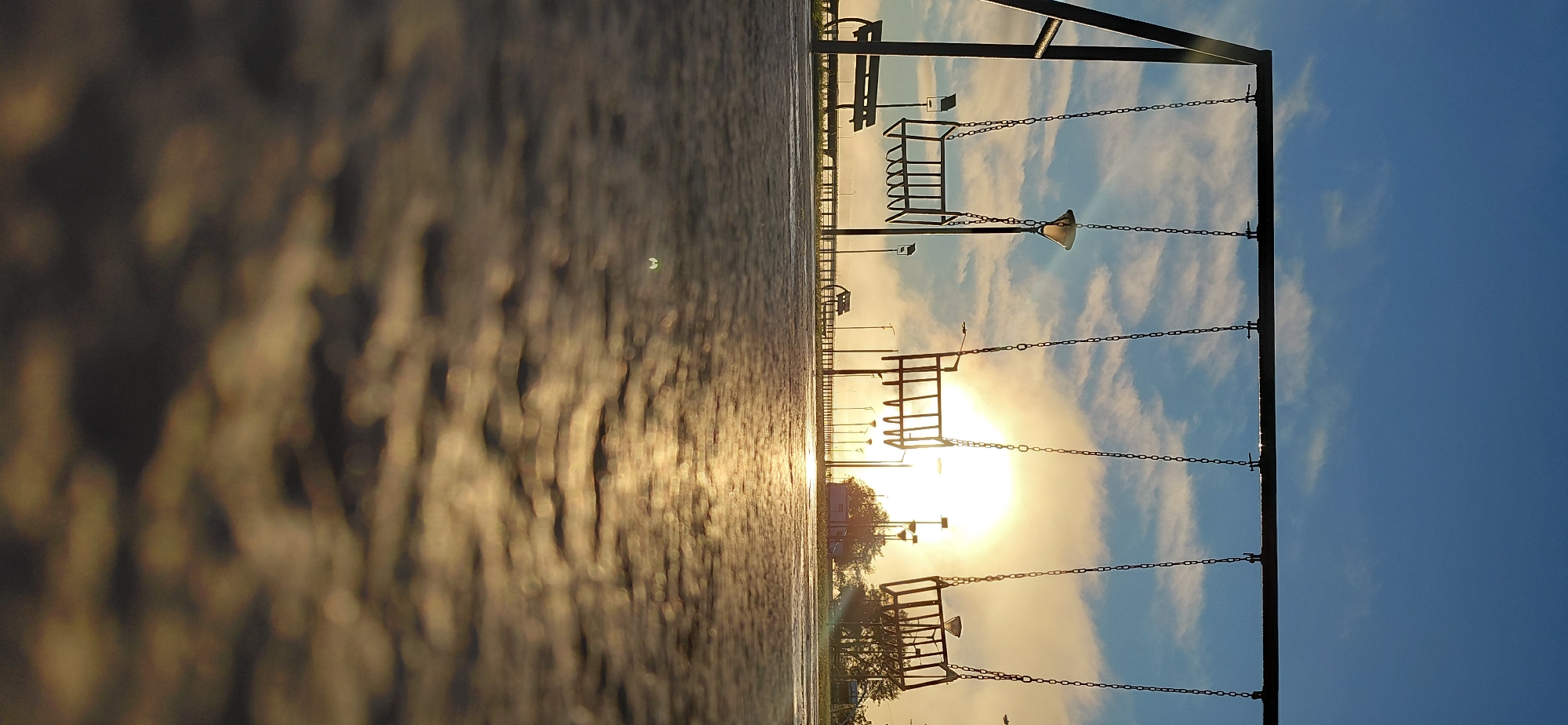 This image captures an early morning scene at a playground, likely located along the waterfront promenade of Encarnación, Paraguay, given the clear sky and the presence of a street lamp. Three empty swings hang from a black metal frame, silhouetted against a bright, hazy sun rising through a layer of clouds. The ground in the foreground is a textured, wet surface, reflecting the golden light of the sun, creating a blurred, abstract pattern. In the background, a bench is visible to the left, and a line of street lamps stretches into the distance, suggesting a well-maintained public space. The overall atmosphere is tranquil and serene, hinting at the start of a new day.